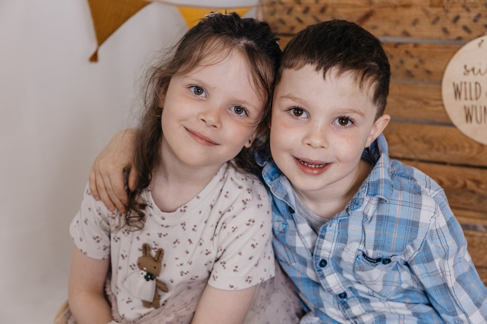 Zwei Kinder sitzen und halten sich freundschaftlich im Arm. Der Hintergrund ist mit Holzelementen dekoriert. Gefertigt von Patricia Bachmann - Kindergartenfotografin aus Göppingen