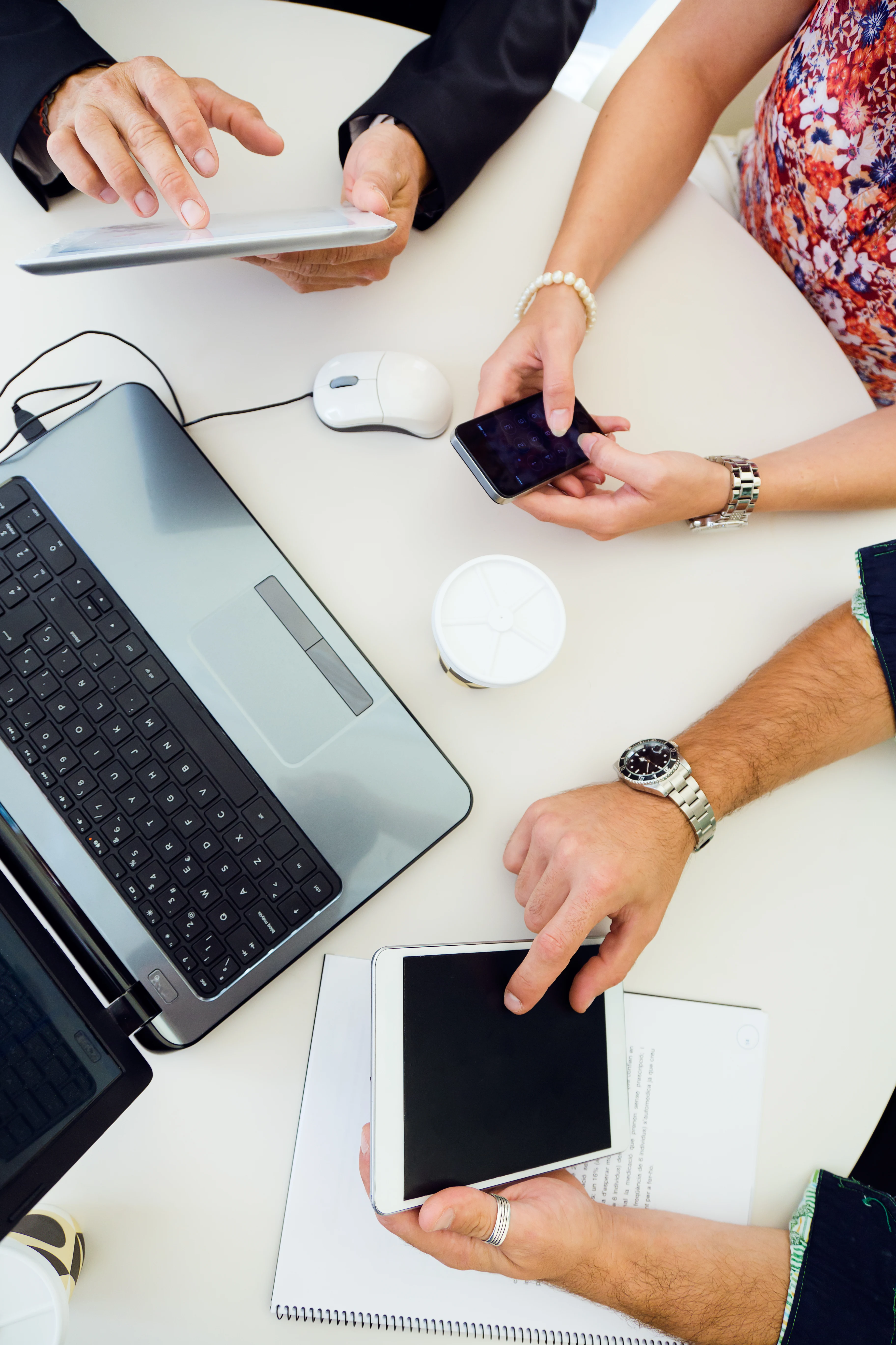 Several hands interacting with technology on a table, including a laptop, tablet, and smartphone, with a notebook and a cup on the surface.