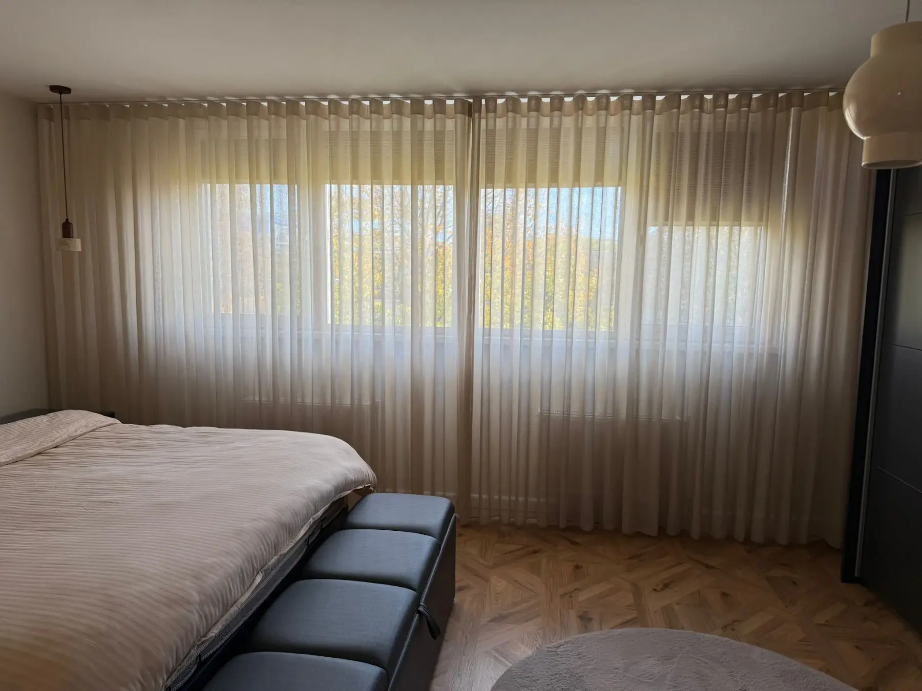 Slaapkamer met een groot bed, een zwarte bank aan het voeteneinde, houten vloer en doorzichtige beige gordijnen voor een breed raam.