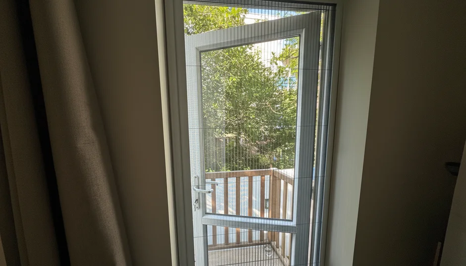 Open deur met een fijne hor naar een balkon met houten balustrade en groene bomen op de achtergrond.