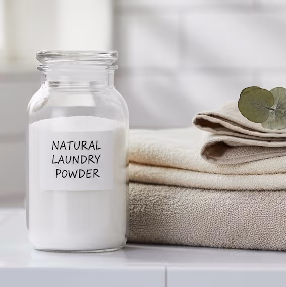 A close-up of a clear glass jar filled with white NATURAL LAUNDRY DETERGENT and labeled with a simple white sticker. The jar sits next to a small stack of three neatly folded neutral-toned towels (beige and light brown), with a dried eucalyptus leaf tucked into the top towel. The background is a white tiled surface.