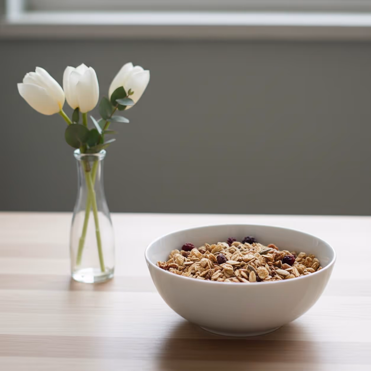 A wide, white ceramic bowl filled with natural cereals (oats, seeds, and dried cranberries) sits on a light wood table. To the left, a small clear glass bud vase holds three white tulips and a sprig of eucalyptus. The background is a plain dark gray wall with a horizontal window above.