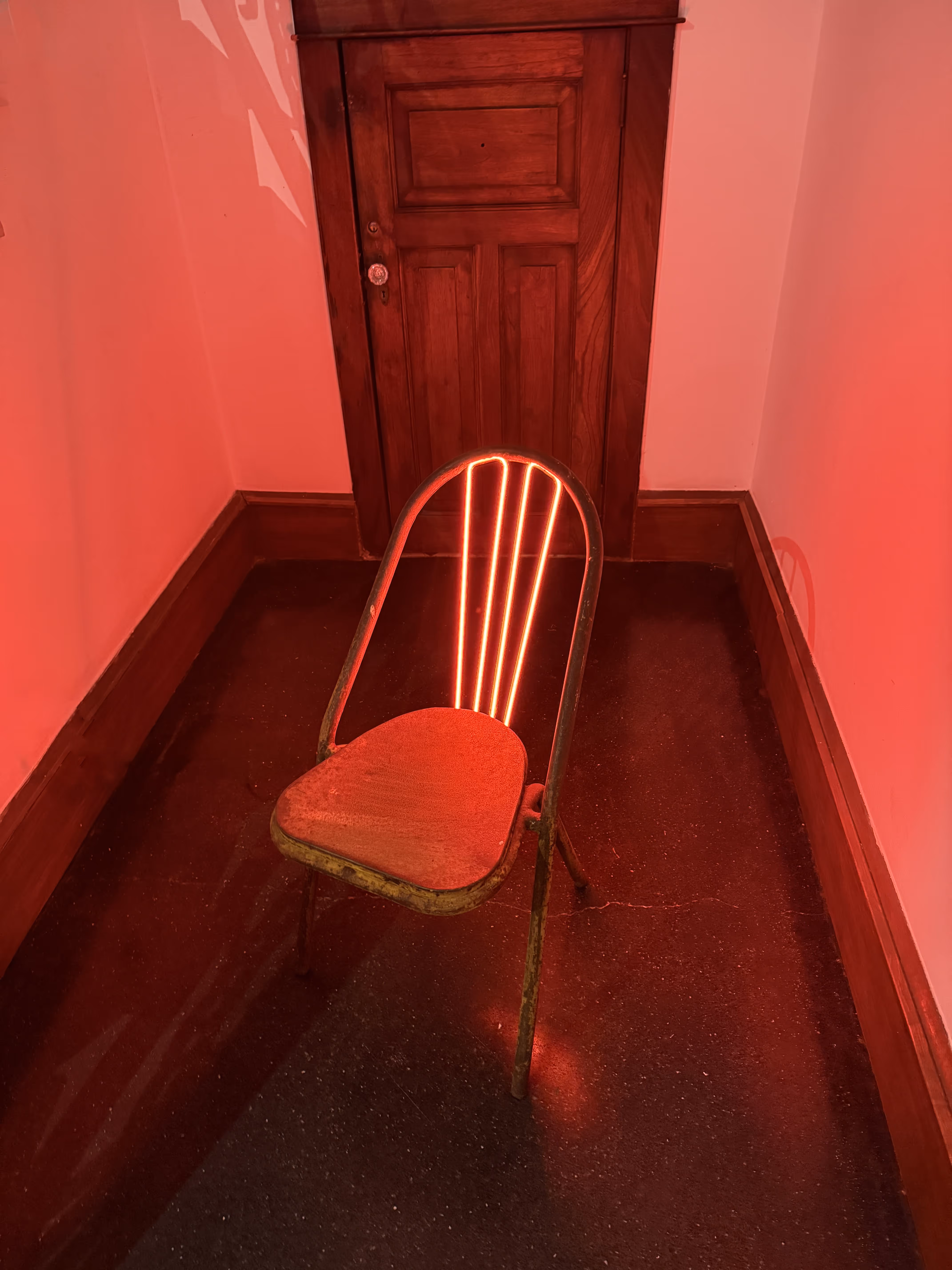 Rusty metal chair with glowing red neon lights on the backrest, placed in a dimly lit corner with a wooden door behind.