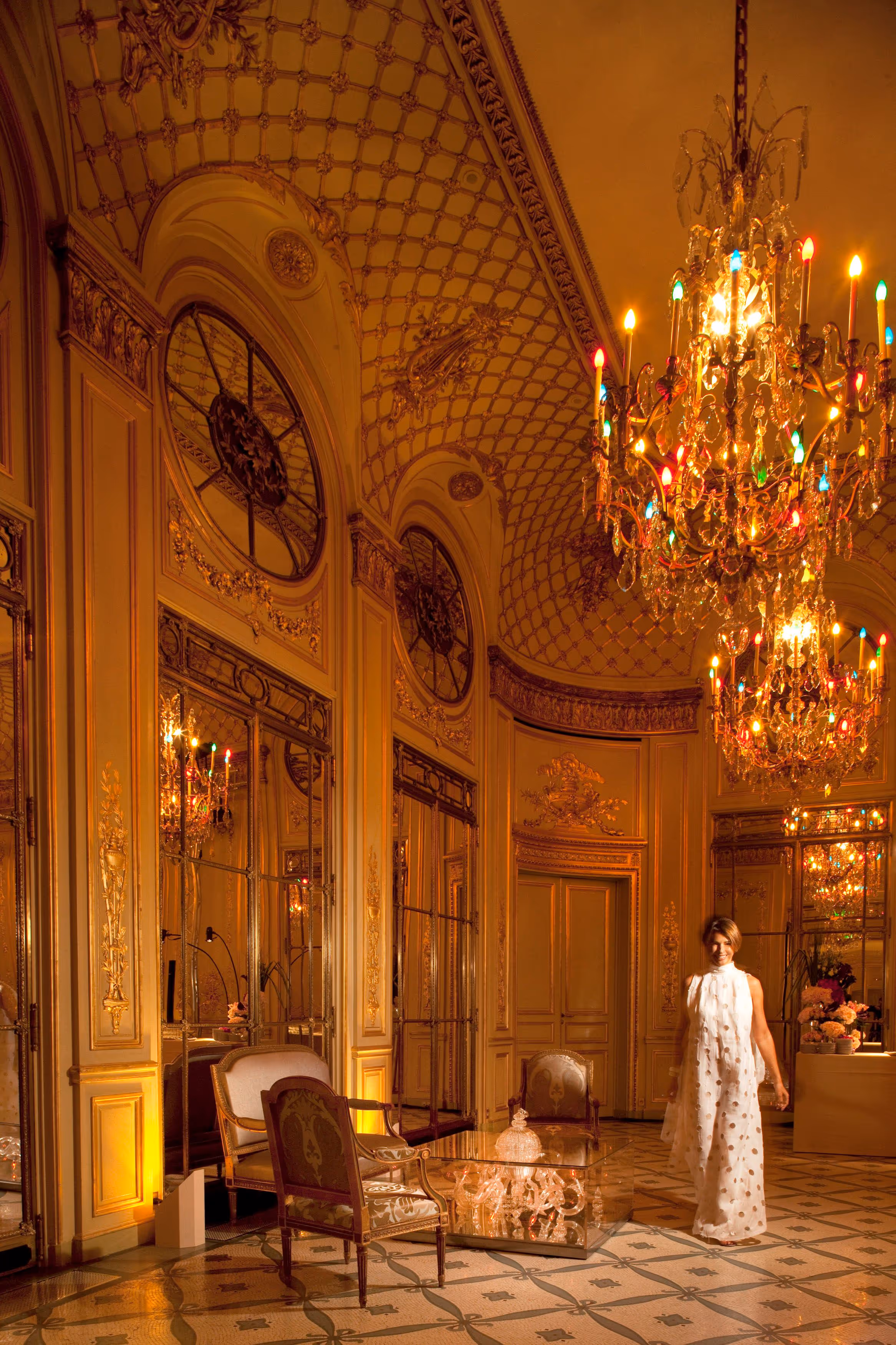 Woman in a white sleeveless dress standing in an ornate room with golden walls, large mirrors, patterned floor, and colorful chandeliers.