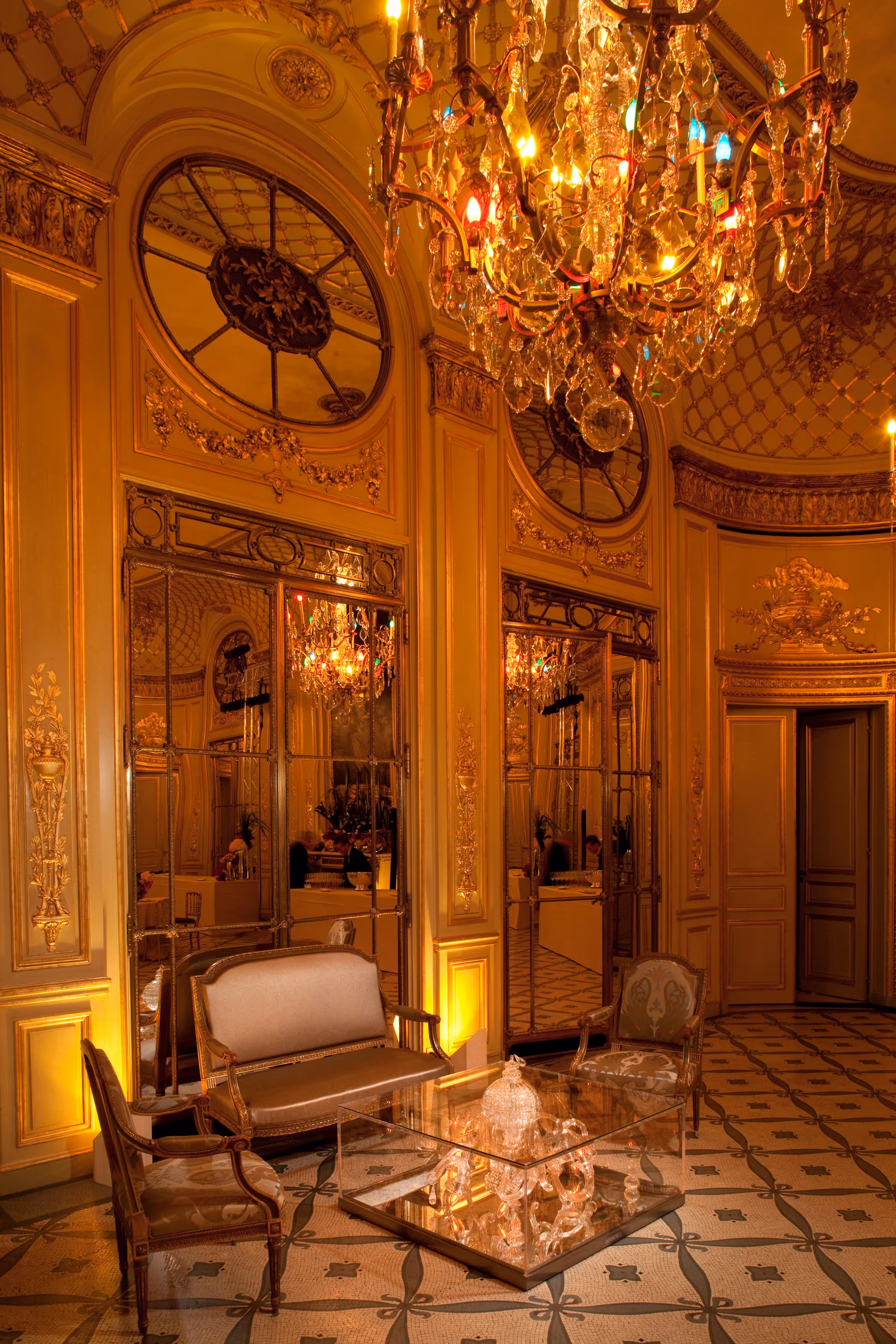 Elegant vintage sitting room with ornate gold walls, mirrored panels, chandelier, and classic furniture surrounding a glass coffee table.