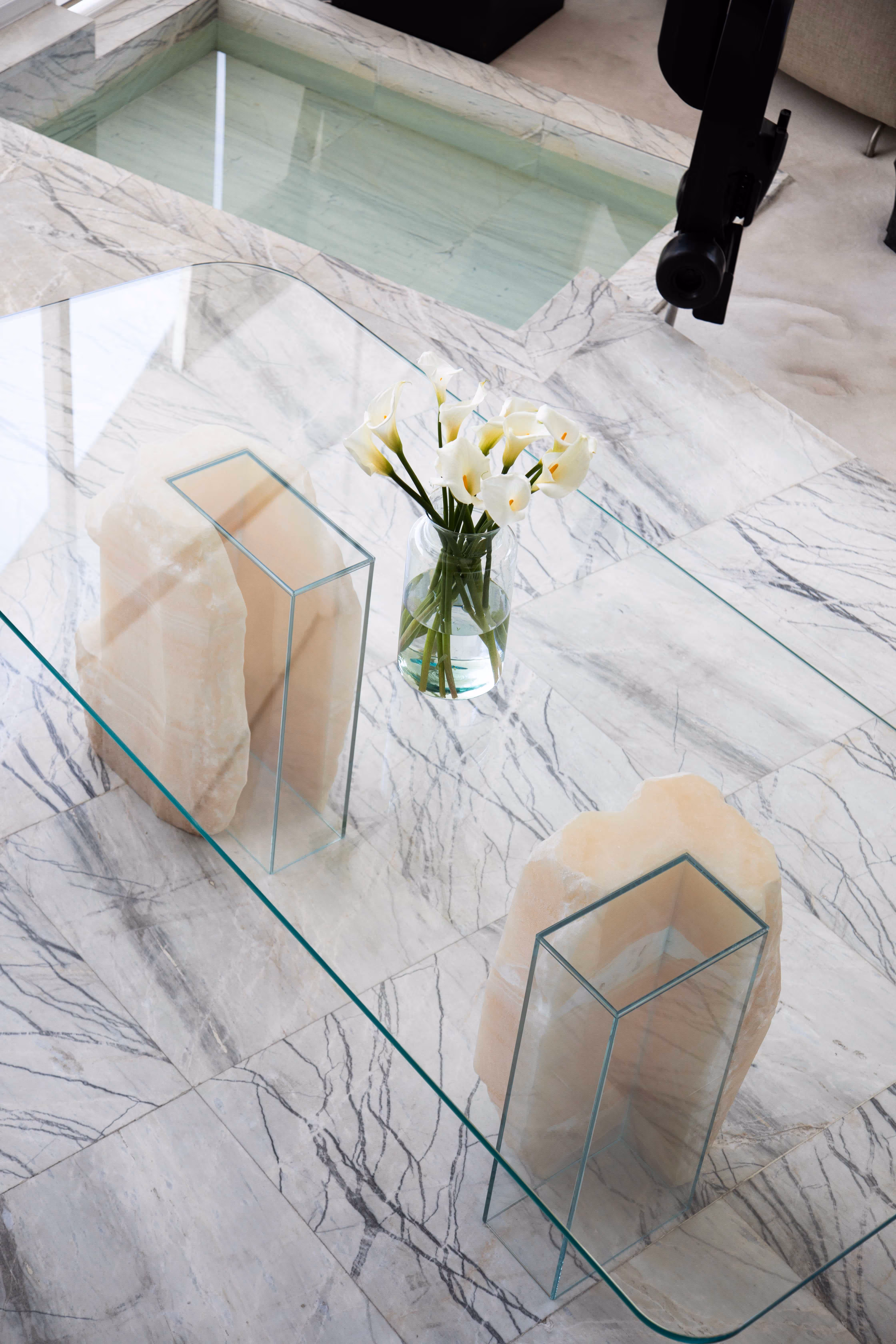 Modern glass coffee table with two stone bases and a vase of white calla lilies on a marble floor near a small indoor pool.