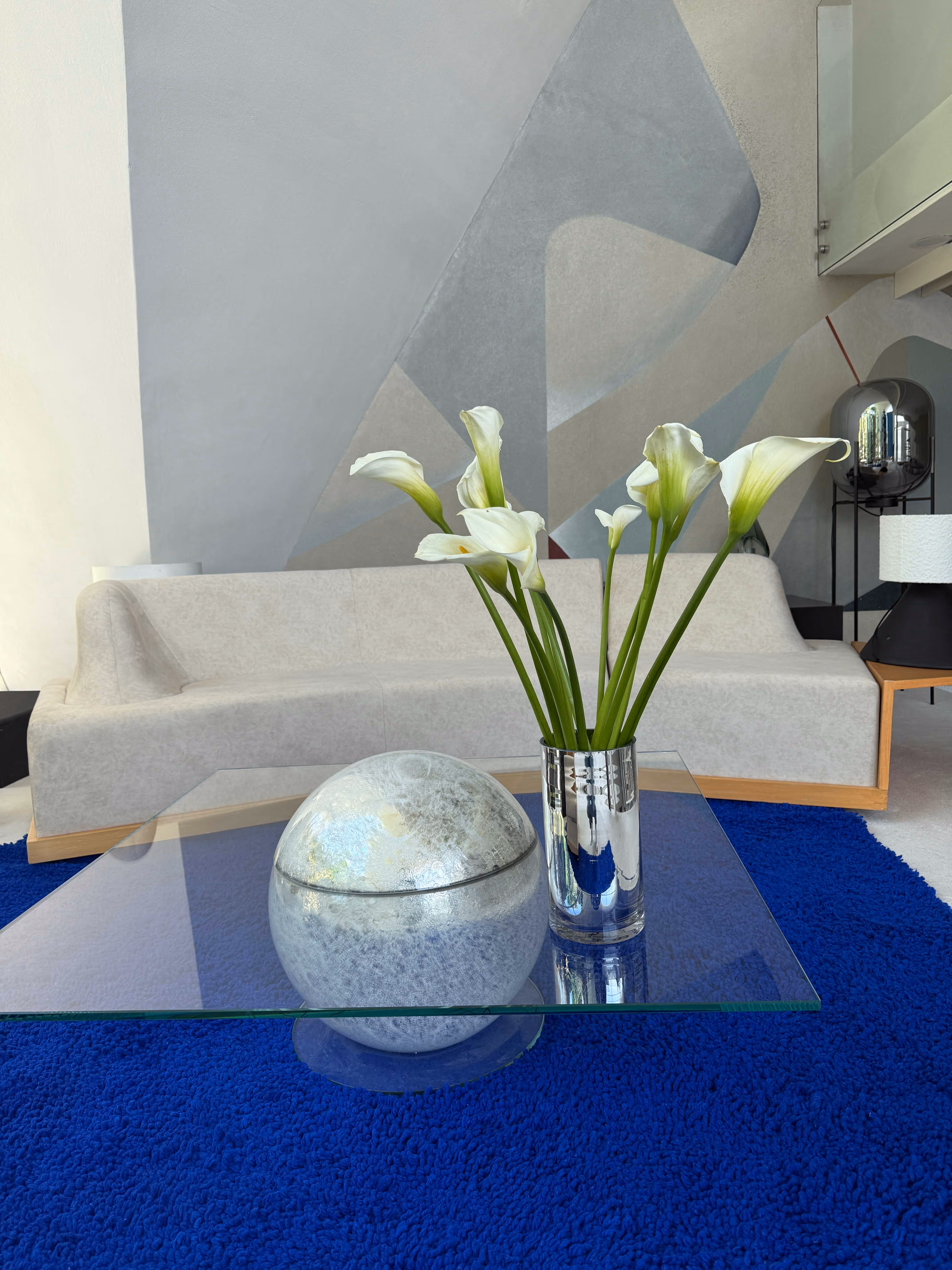 Modern living room with a white sofa, abstract gray wall art, blue shag rug, and a glass coffee table holding a silver spherical sculpture and a vase with white calla lilies.
