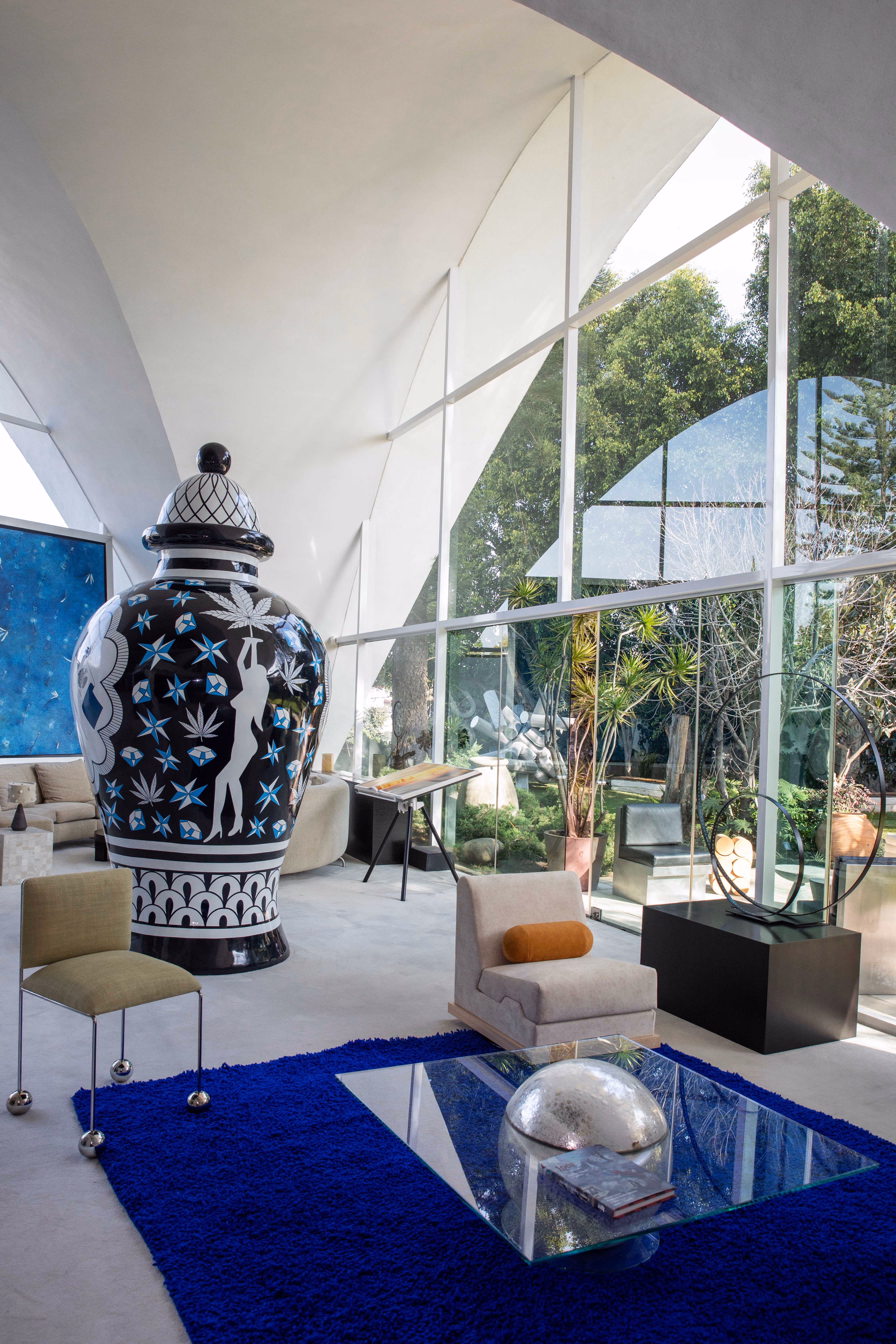 Modern living room with white curved ceiling, large black-and-white decorative vase, blue rug, glass coffee table, and floor-to-ceiling windows overlooking garden.