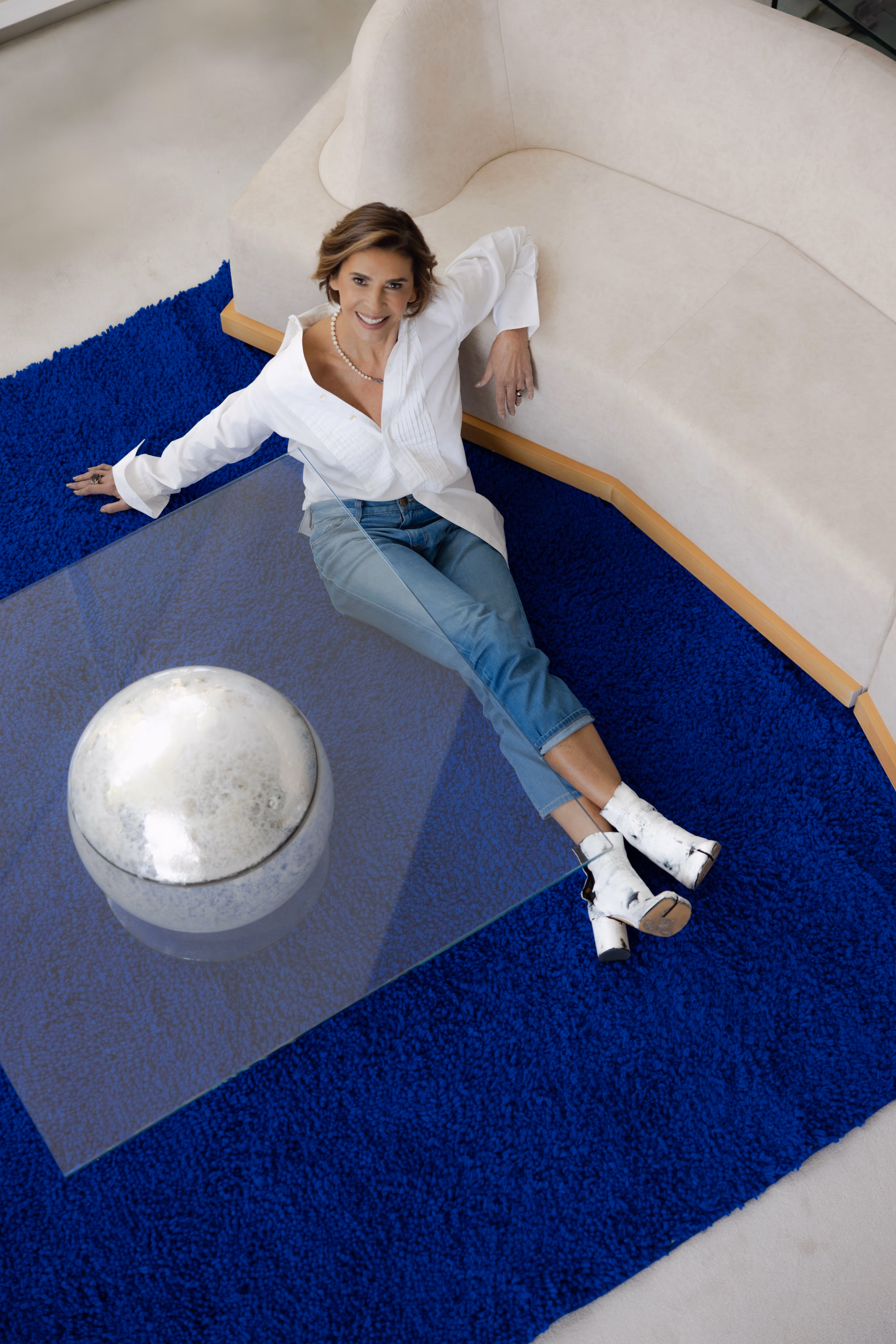 Woman in a white blouse and blue jeans reclining on a blue shag rug beside a beige curved sofa and glass coffee table with a reflective silver orb.