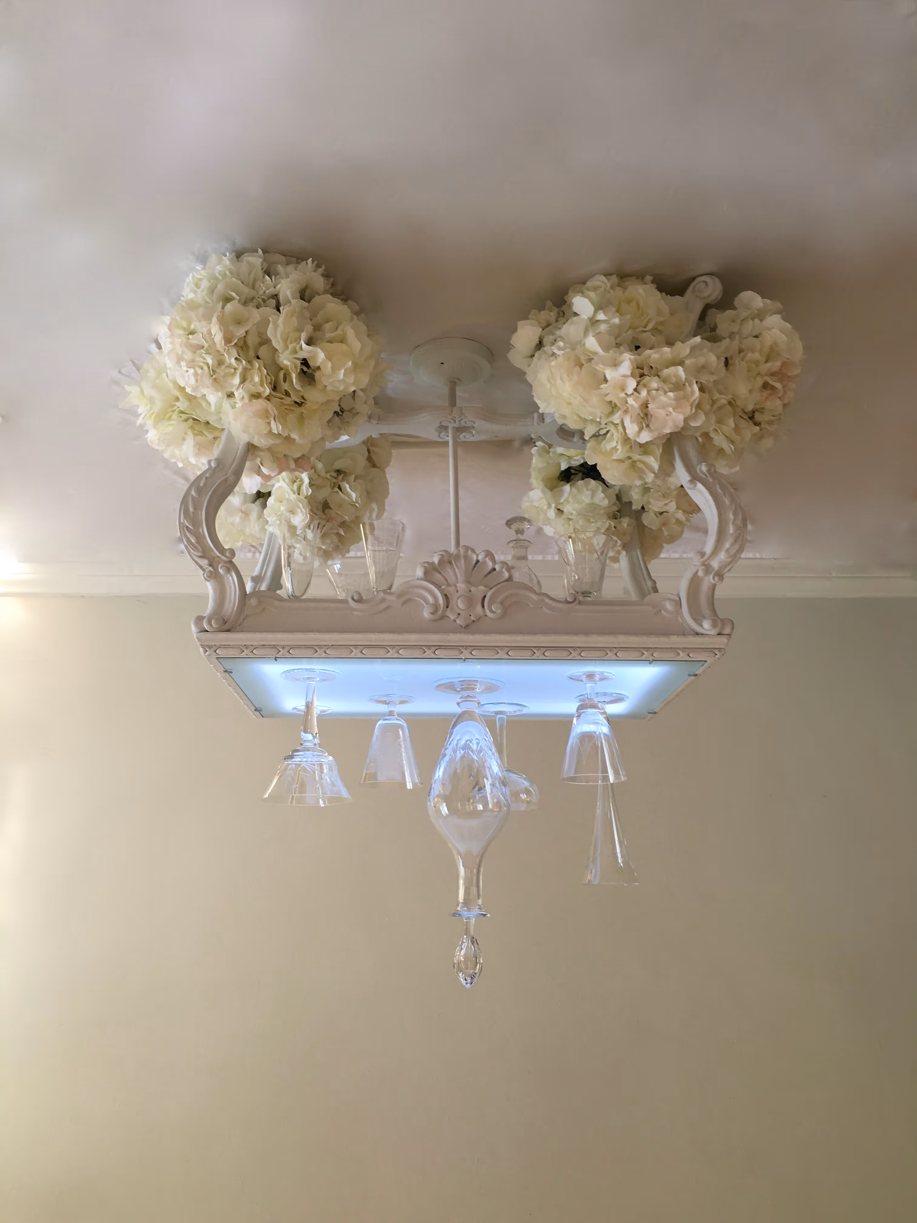 Ceiling light fixture decorated with white hydrangea flowers and hanging upside-down glassware.