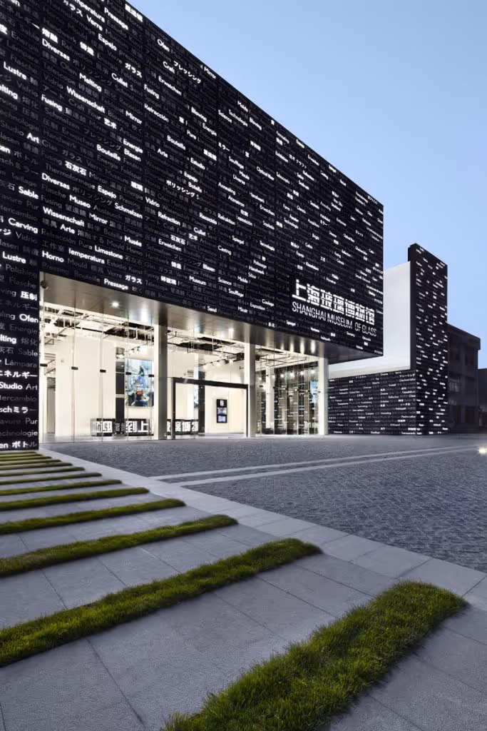 Modern black building of the Shanghai Museum of Glass with illuminated text and a spacious entrance, stairs with grass strips in the foreground.