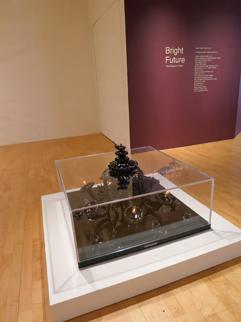A dark, ornate chandelier displayed under a clear protective case on a white pedestal in an art gallery with a purple wall featuring text reading 'Bright Future: New Designs in Glass.'