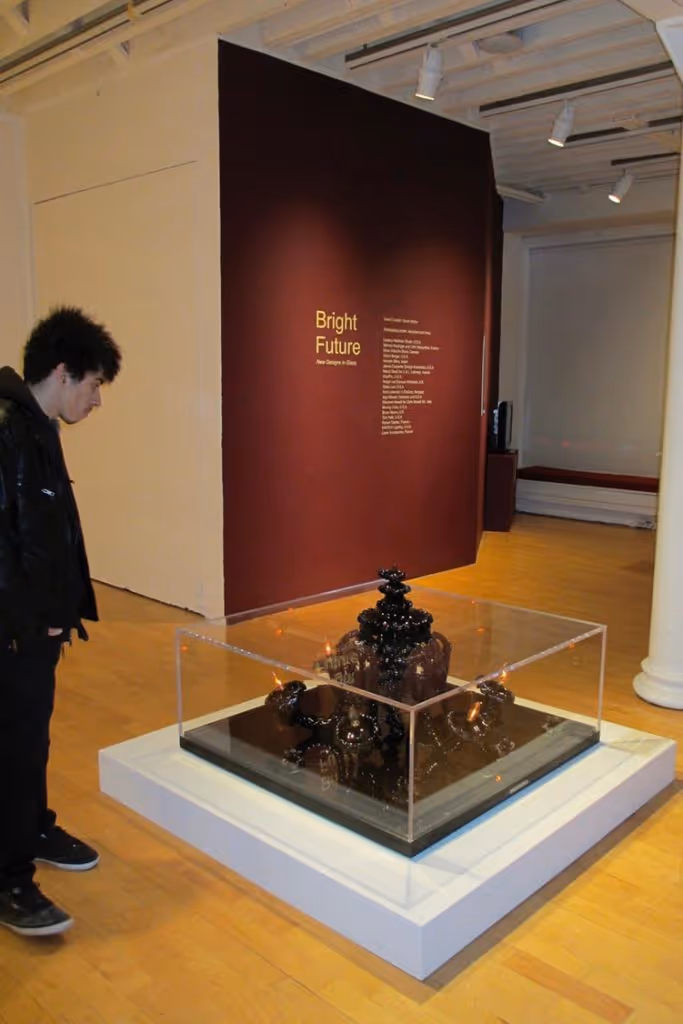 Man in black jacket observing a black sculptural art piece inside a transparent display case in an art gallery with a maroon wall titled 'Bright Future'.