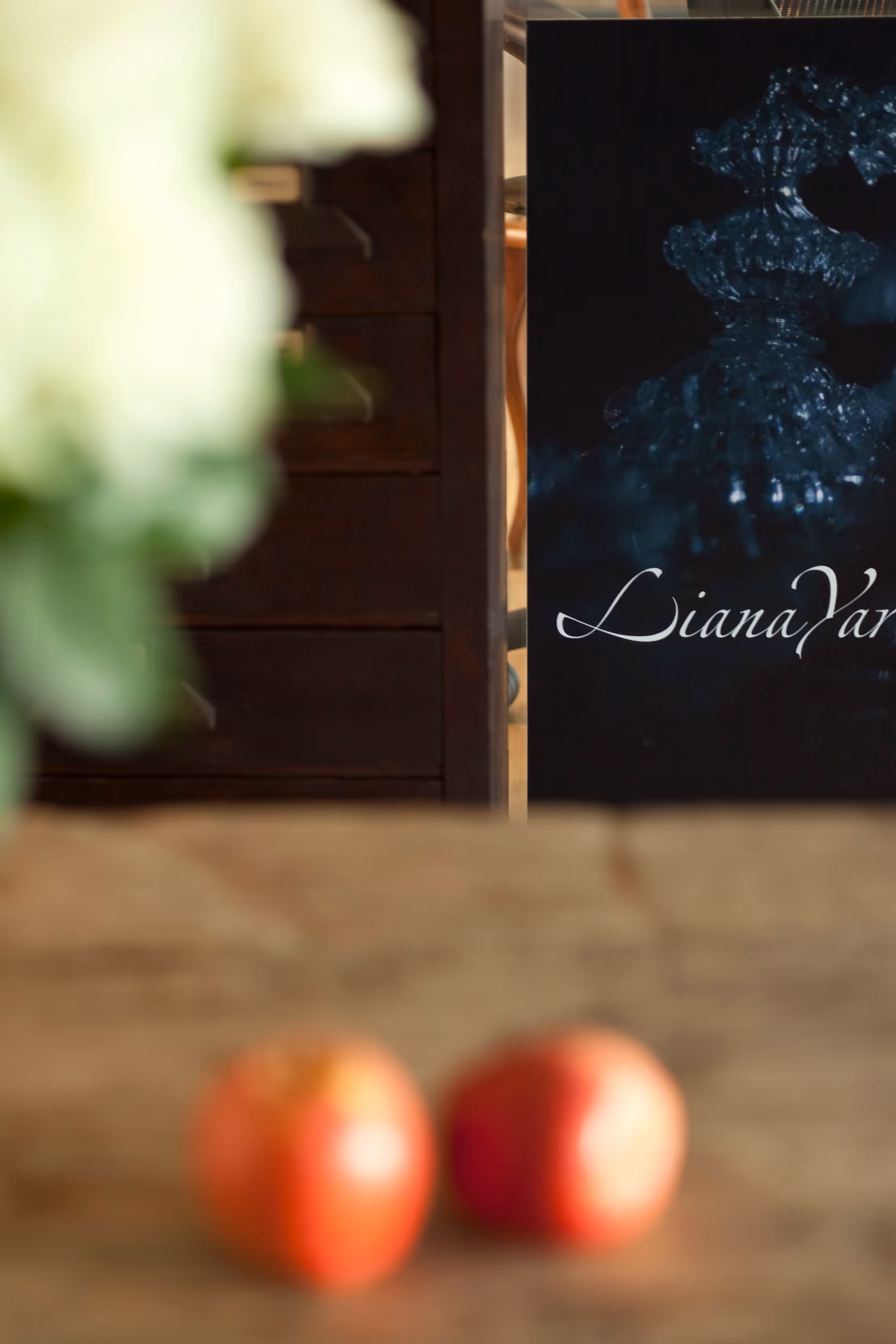 Two out-of-focus apples on a wooden table with a dark sign in the background reading 'LianaYar' and part of a white flower arrangement on the left.