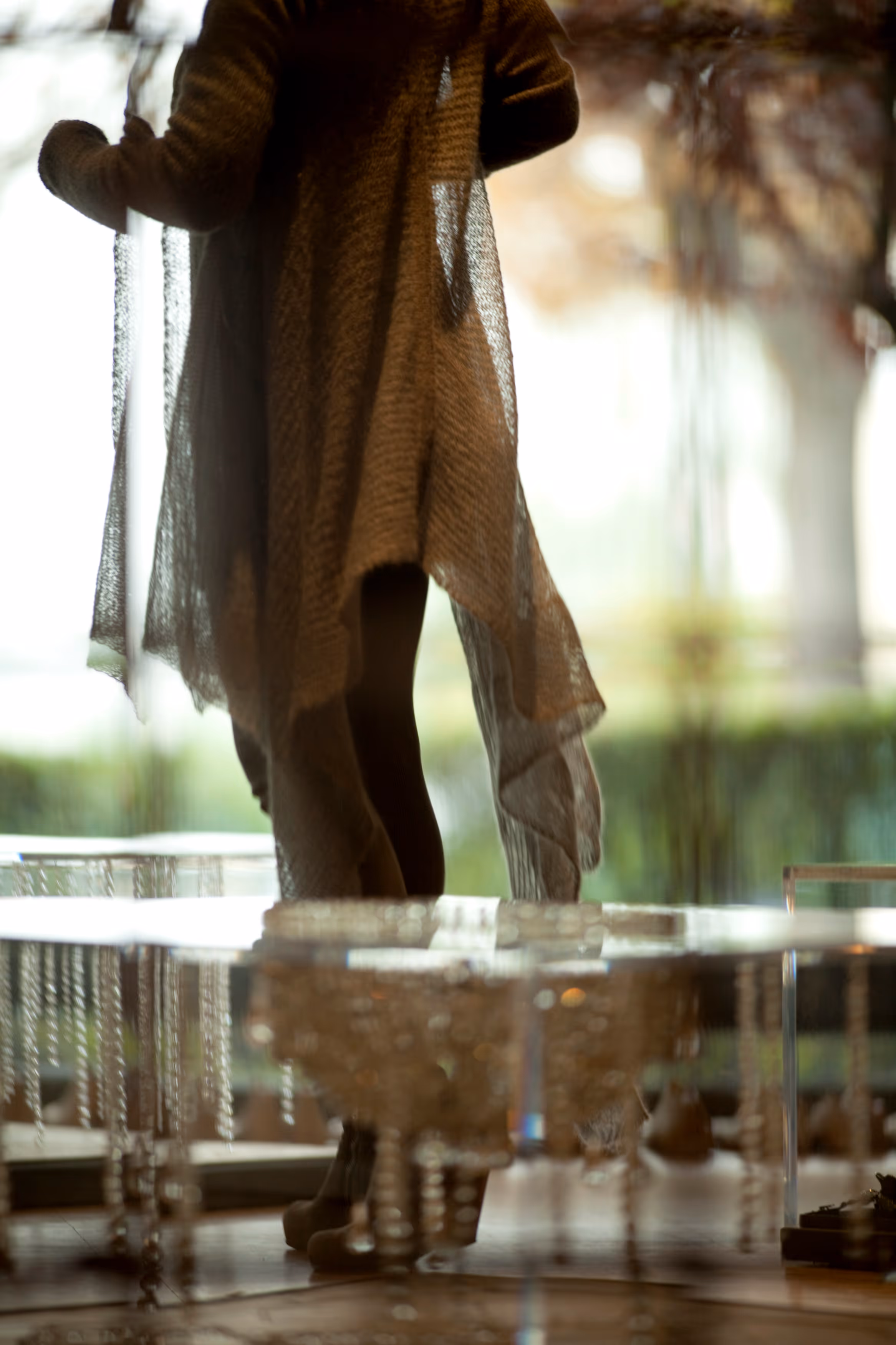 Lower part of a person wearing a brown knitted shawl and black leggings standing indoors near transparent decor.