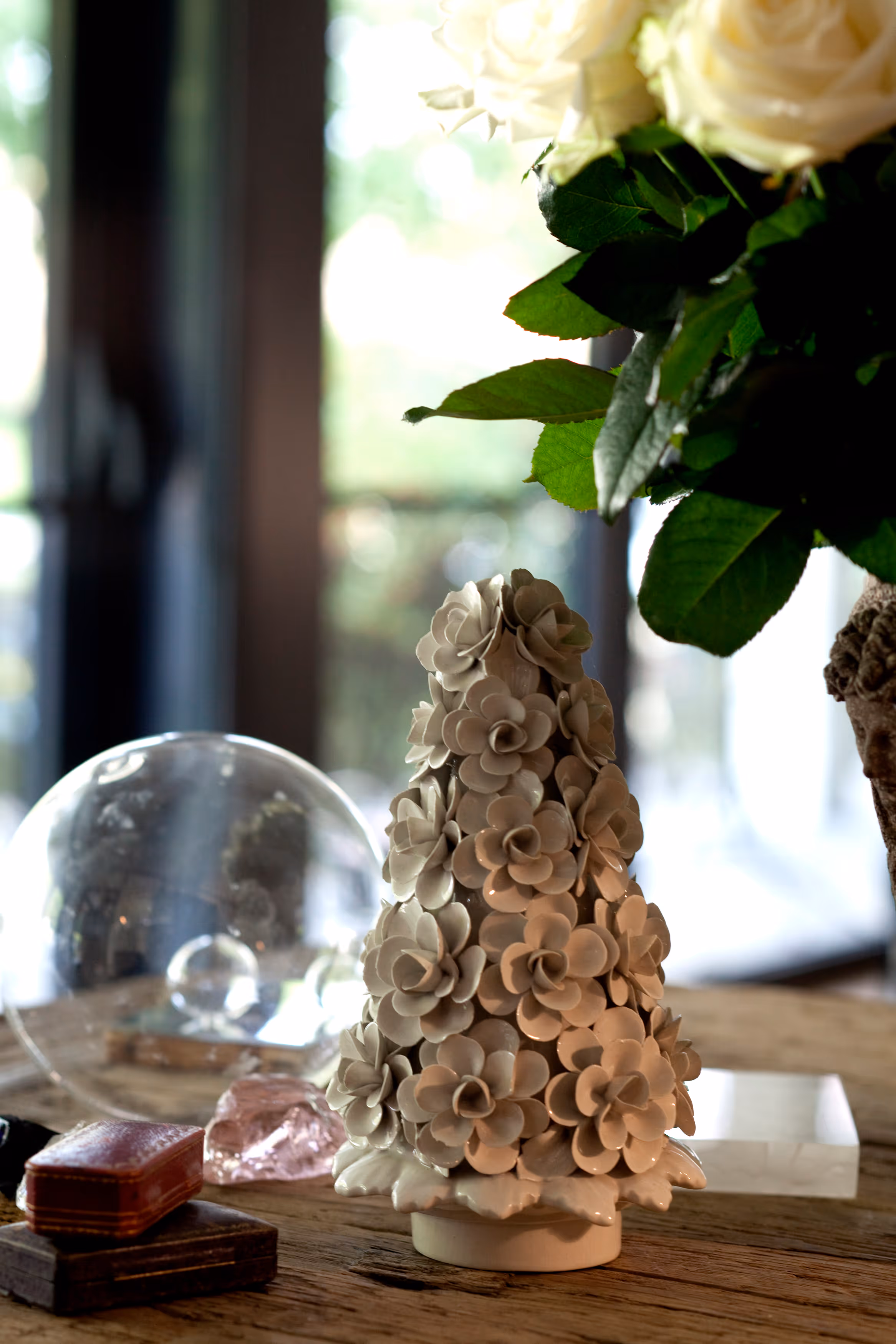 White ceramic decorative tree covered in flower shapes on a wooden surface with a glass globe and a bouquet of white roses nearby.