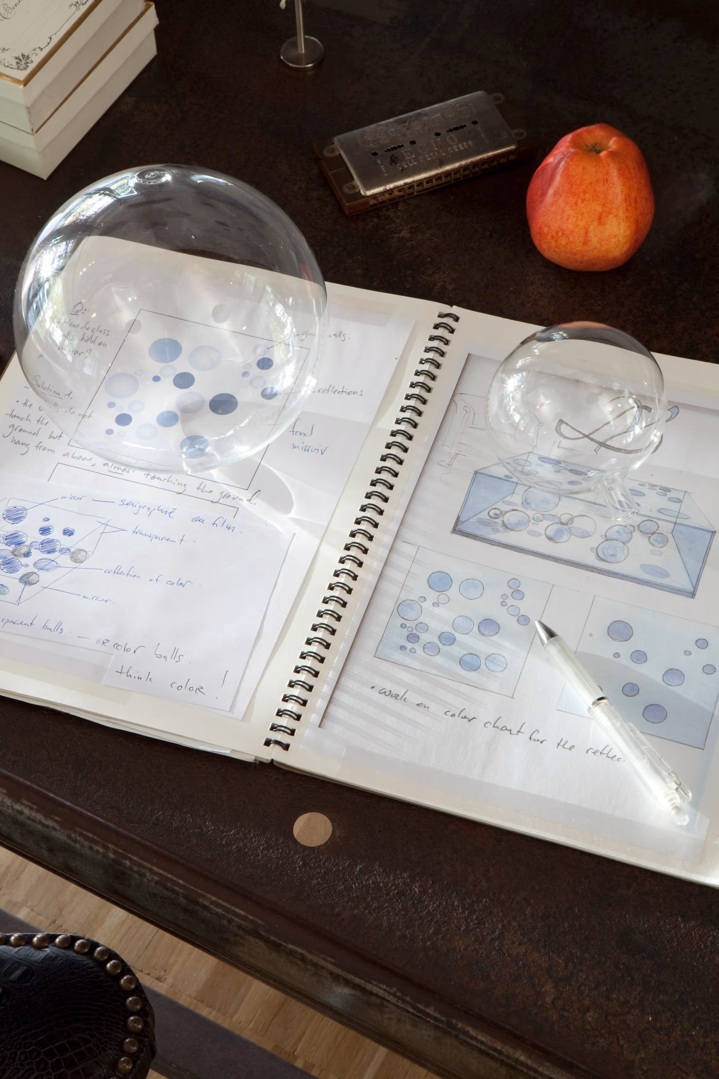 Open sketchbook on a dark wooden table showing bubble design drawings and notes, with two clear glass spheres, a white pen, a red apple, and a harmonica nearby.