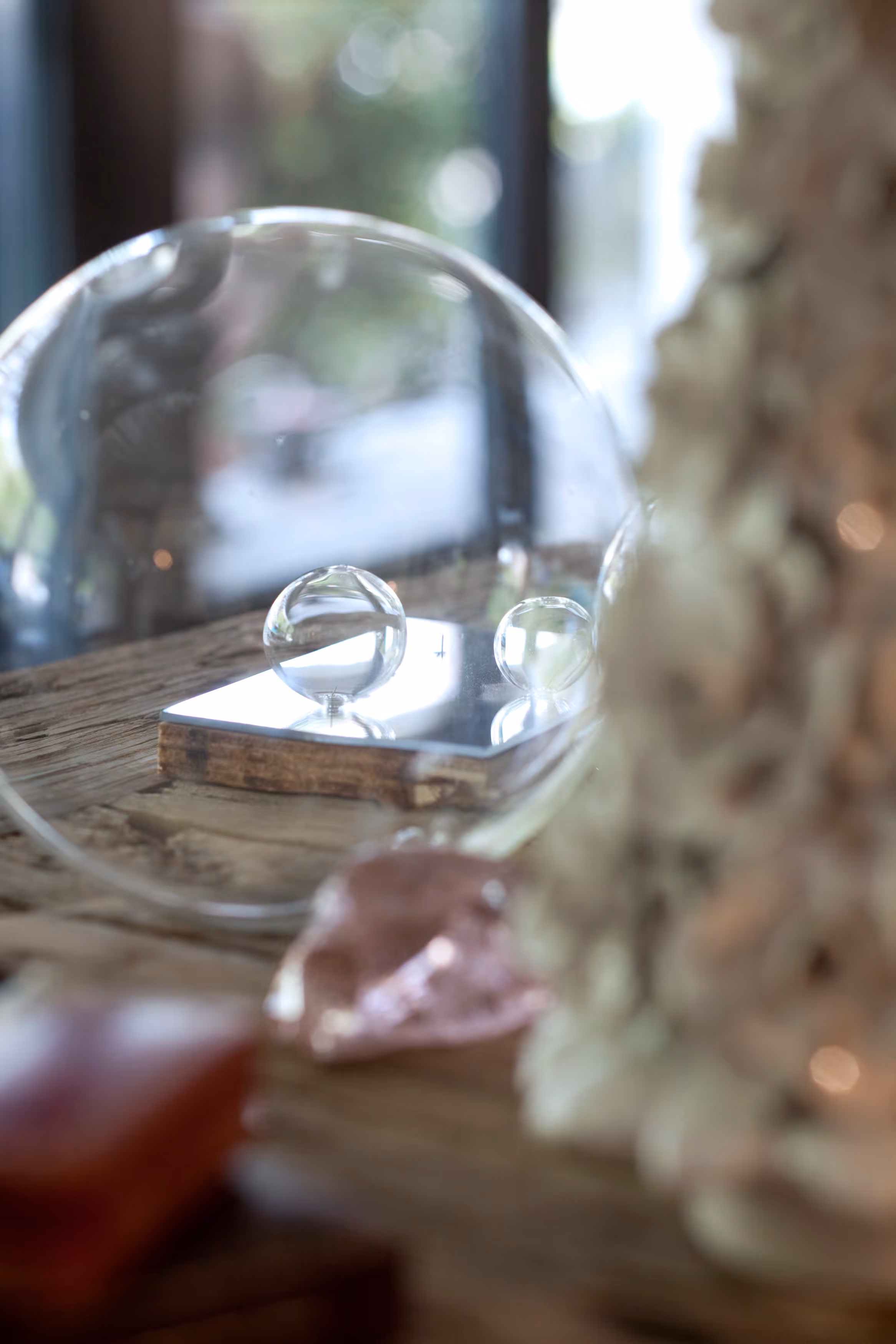 Two clear glass spheres on a small mirrored square placed on a wooden surface, with blurred objects in the foreground and background.
