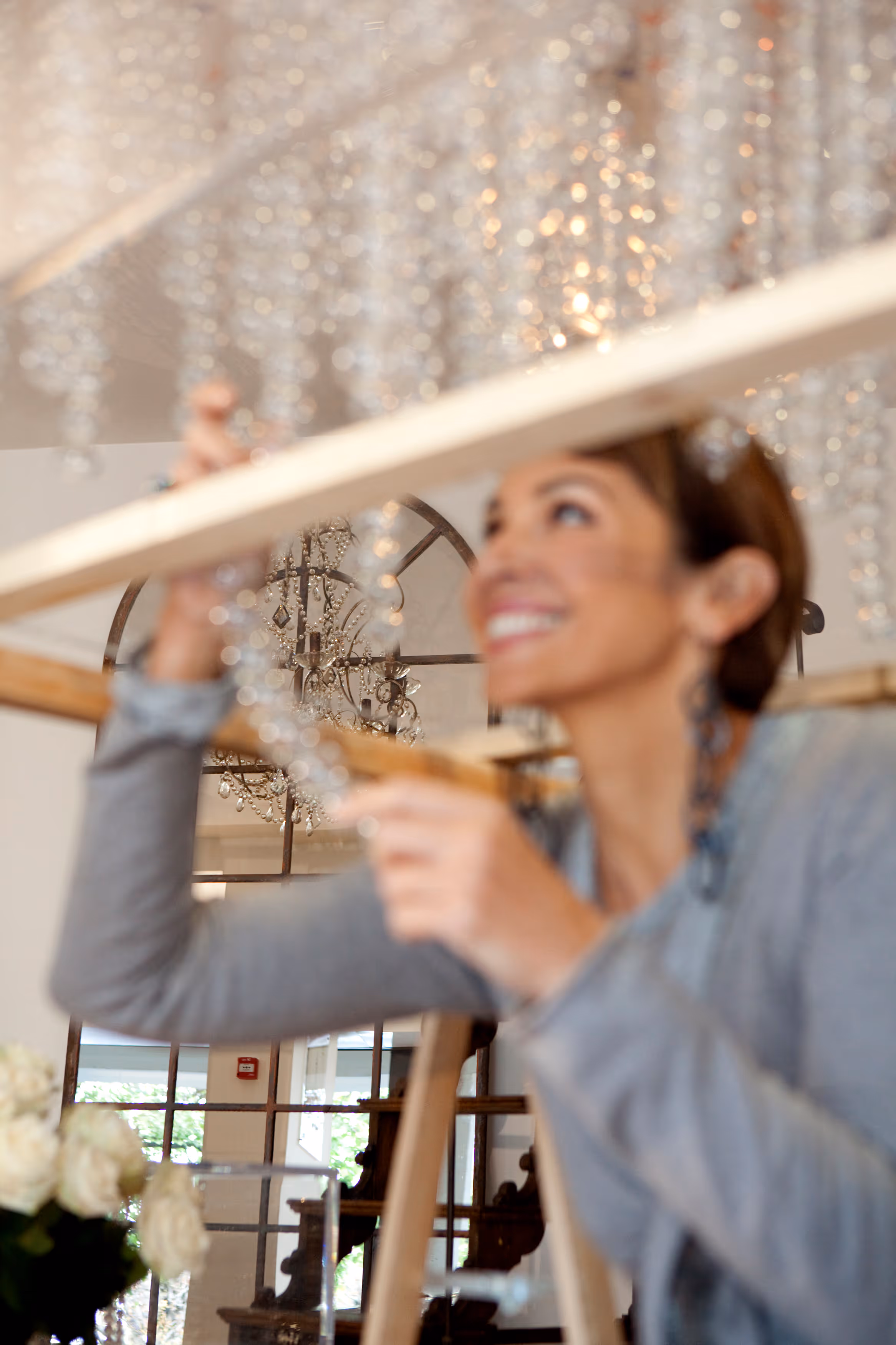 Woman installing or adjusting a crystal chandelier indoors with a window and mirror in the background.
