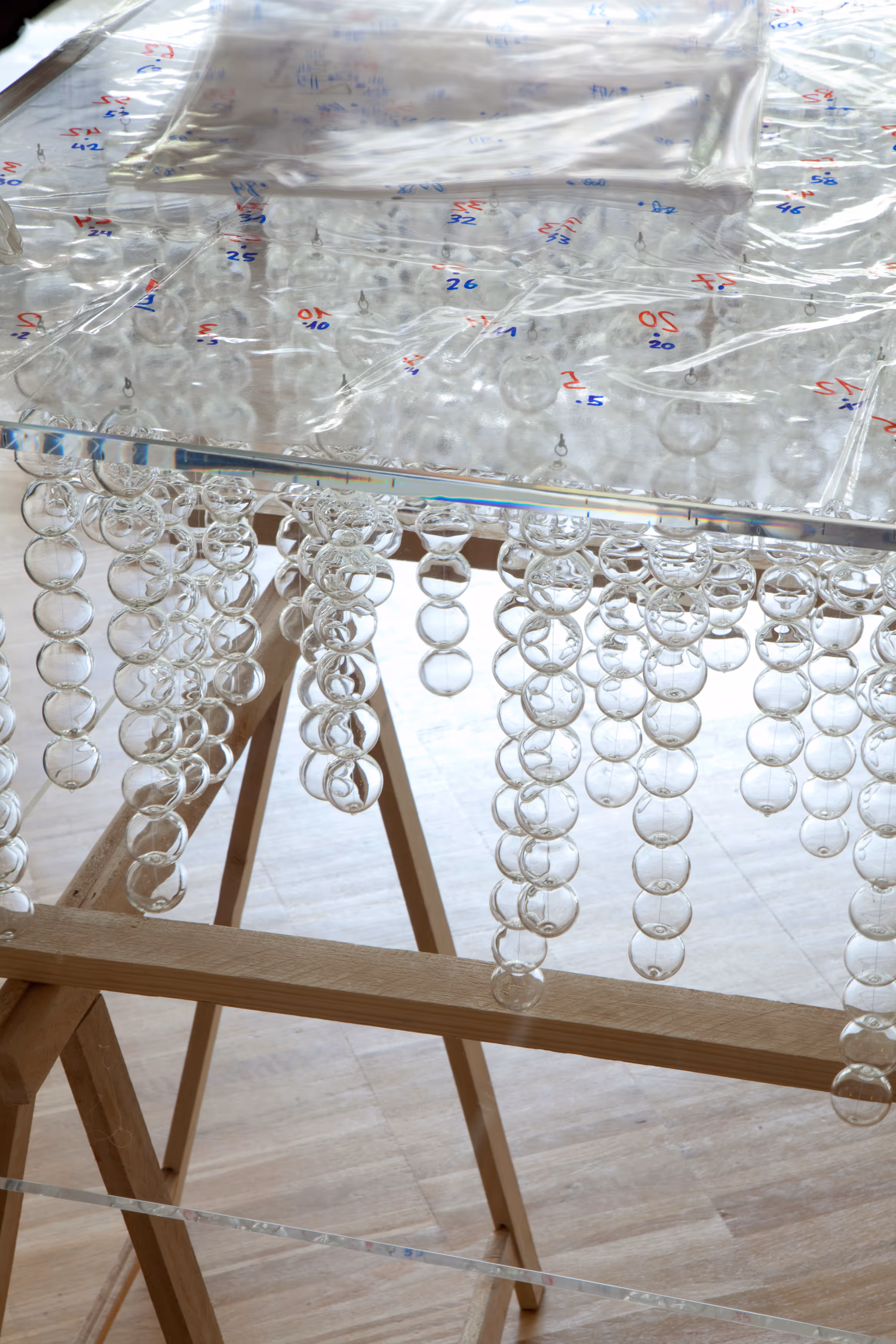 Close-up of a transparent acrylic table with numbered markings and hanging clear glass spheres above a wooden frame on a light floor.