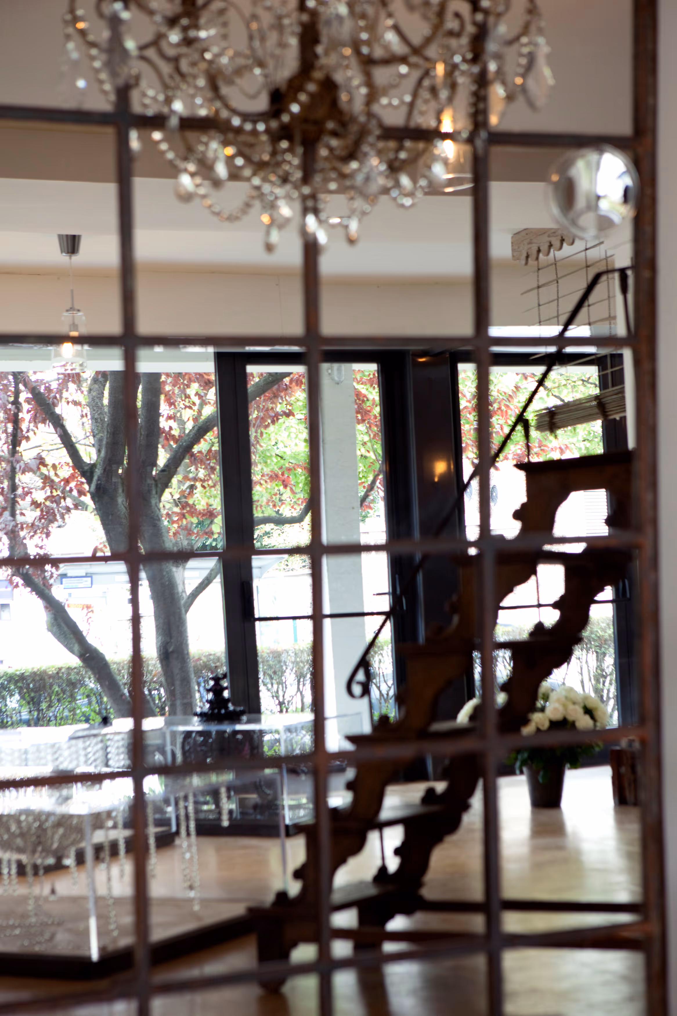 Reflection in a window showing a wooden spiral staircase, chandelier, and room with large windows overlooking trees.