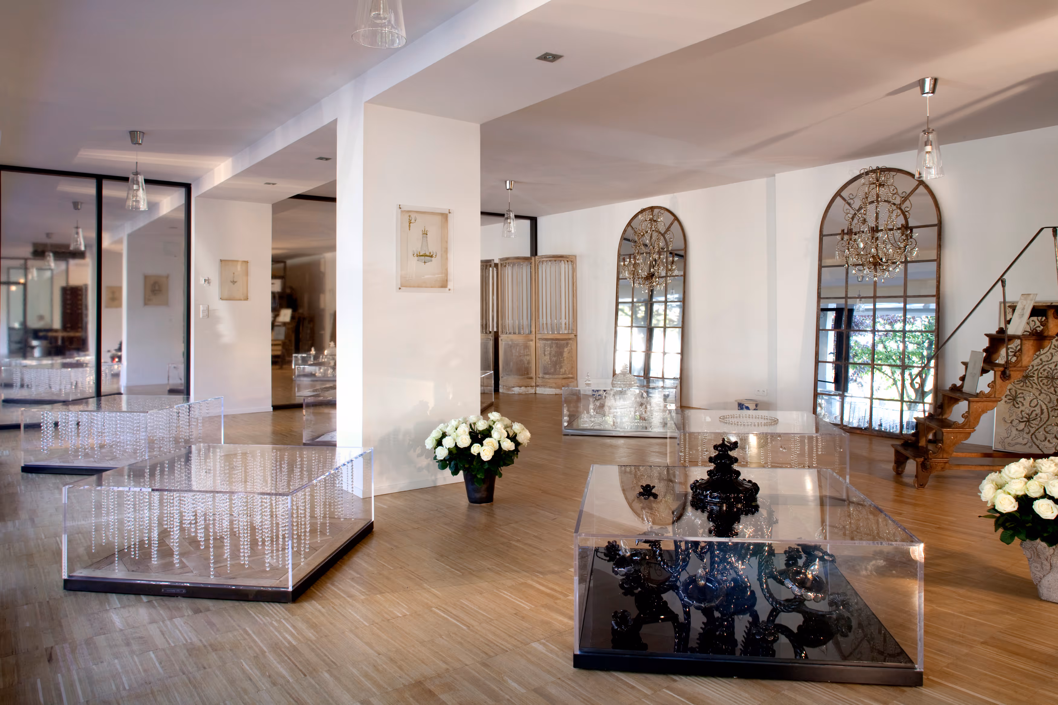 Bright modern gallery room with transparent display cases featuring decorative objects, large mirrors, wooden staircase, and white roses.