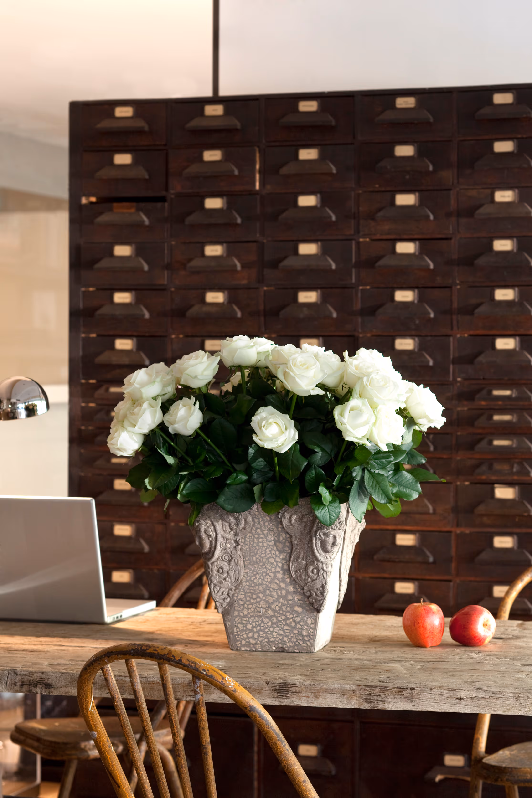 Rustic wooden table with a decorative vase of white roses, two red apples, a laptop, and a metallic desk lamp in front of a wall of dark wooden drawers.