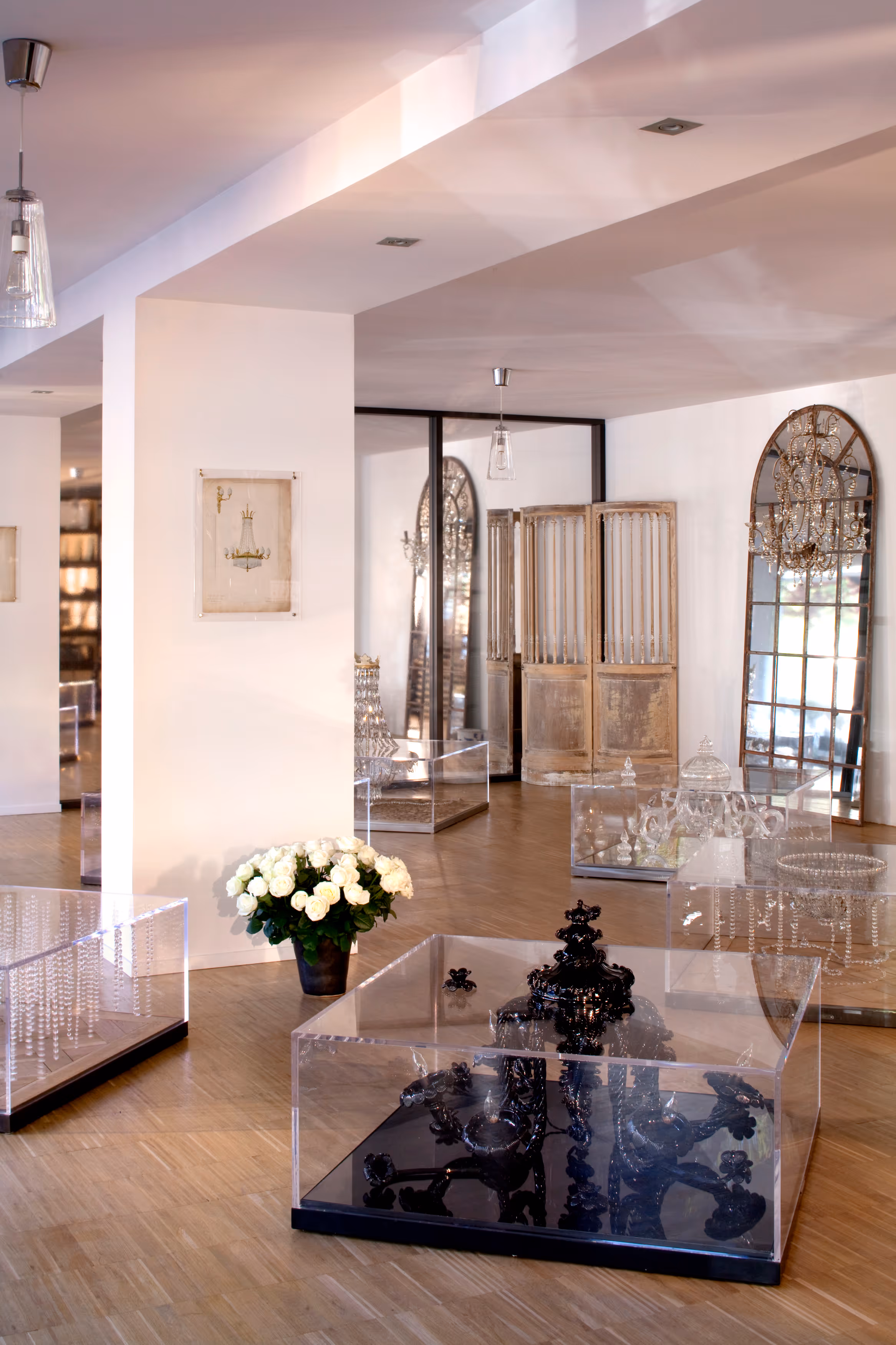 Modern gallery room with wooden flooring displaying chandeliers and decorative glass pieces in transparent cases, a pot of white roses, and large ornate mirrors.