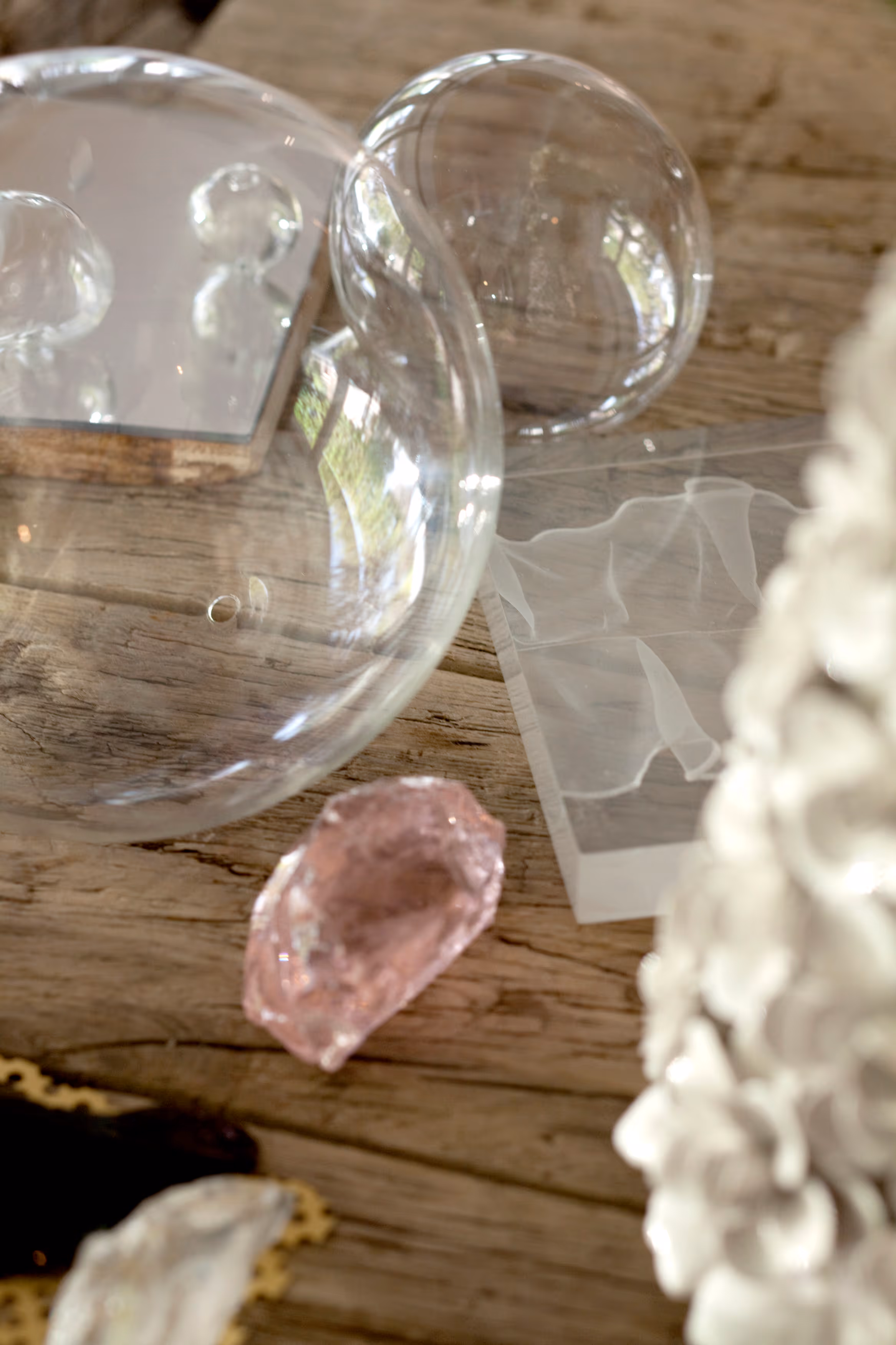 Close-up of transparent glass bubbles, a pink crystal, and white floral decoration on a wooden surface.