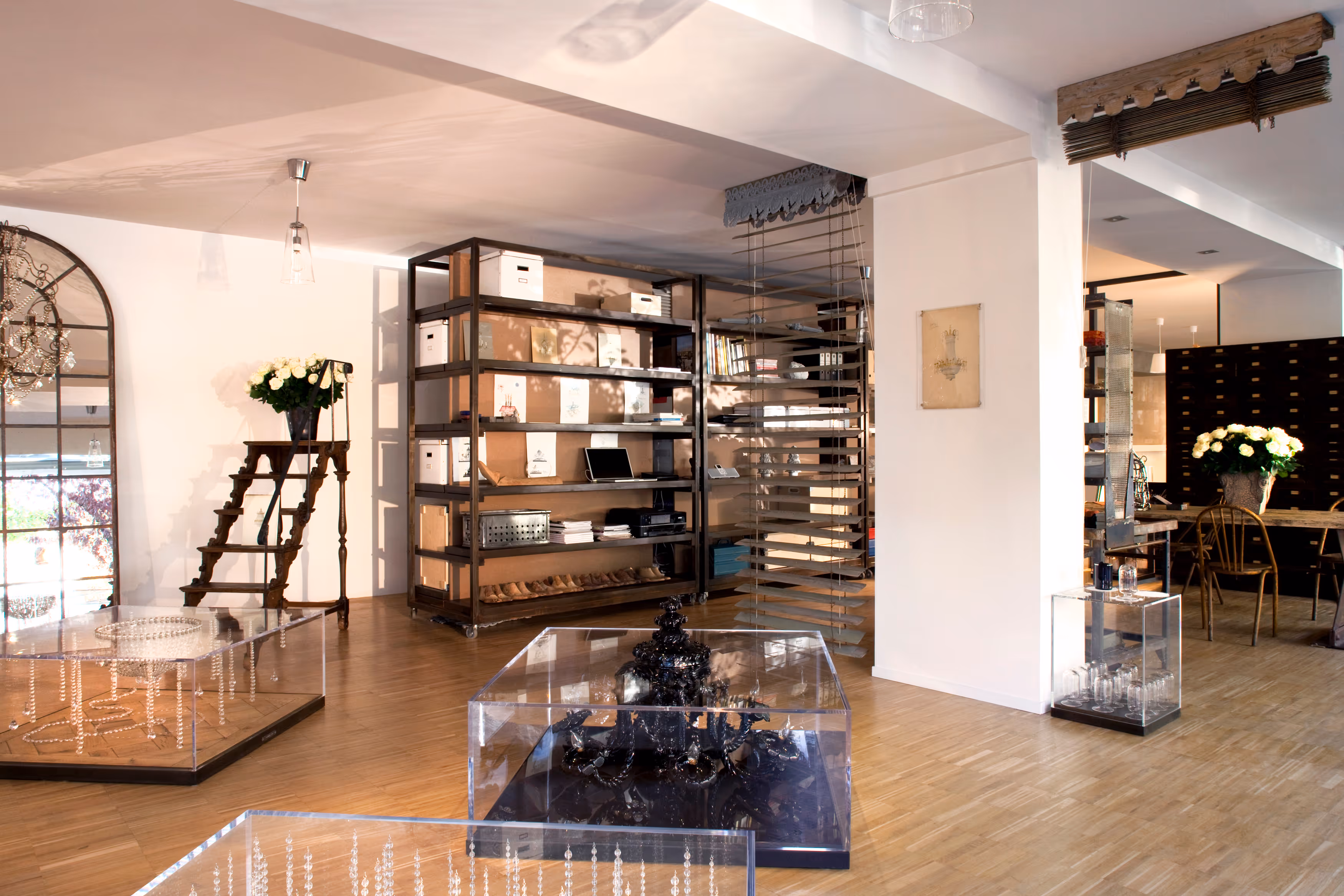 Modern interior with wooden floor, large shelving unit, clear display cases with chandeliers, and floral arrangements.
