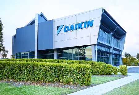 Modern Daikin office building with large glass windows surrounded by green hedges and a sidewalk.
