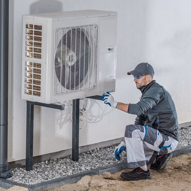 Technician wearing gloves and safety glasses kneeling while inspecting an outdoor air conditioning unit mounted on a wall.