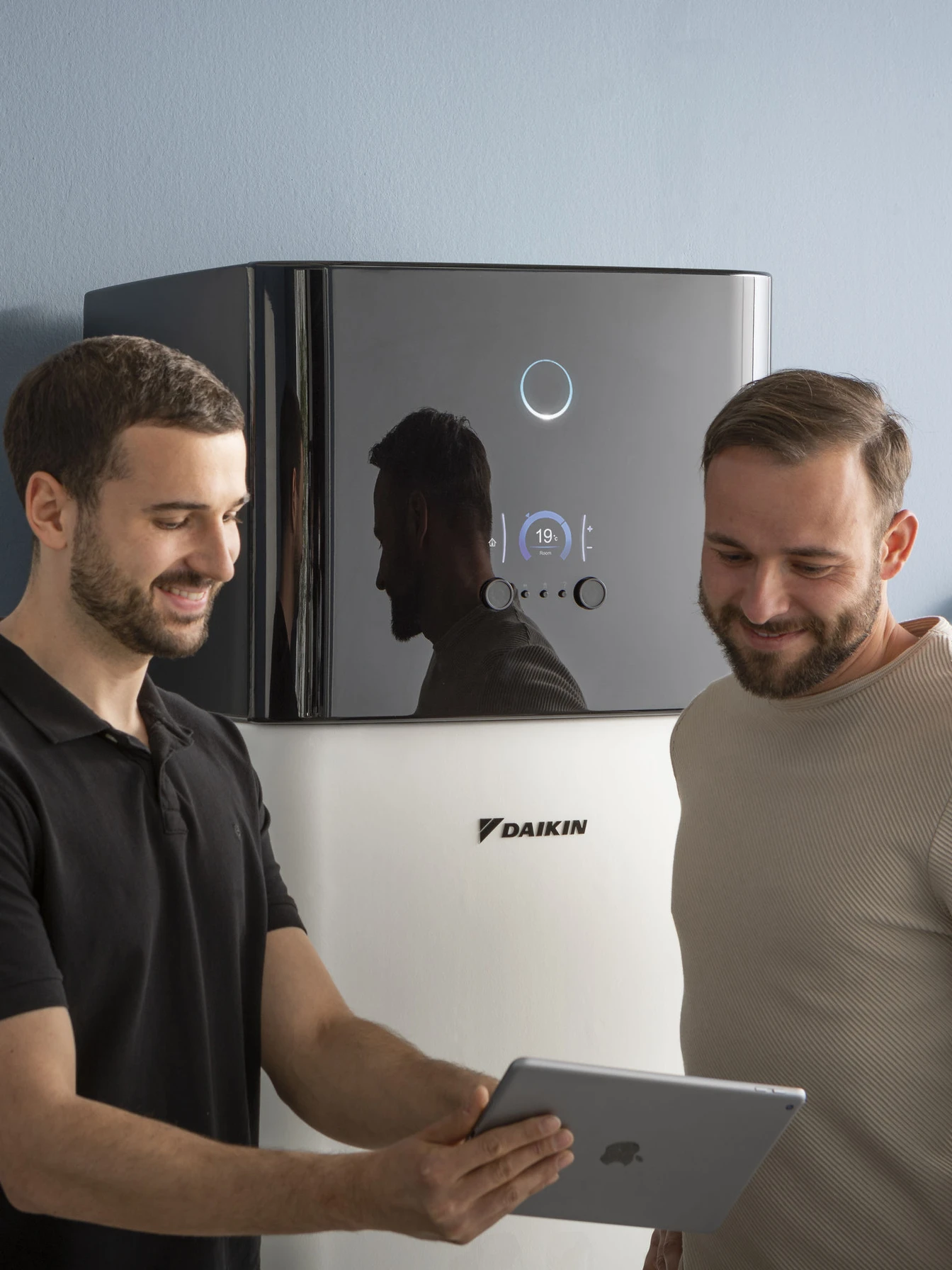 Two men smiling while looking at a tablet in front of a Daikin heating unit with a digital display showing 19 degrees Celsius.