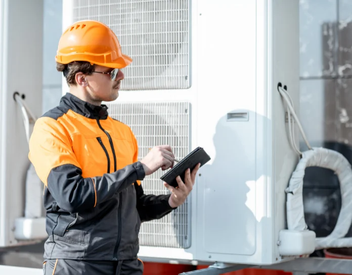 Technician in orange and black work uniform with hard hat inspecting an outdoor heat pump unit while using a tablet.