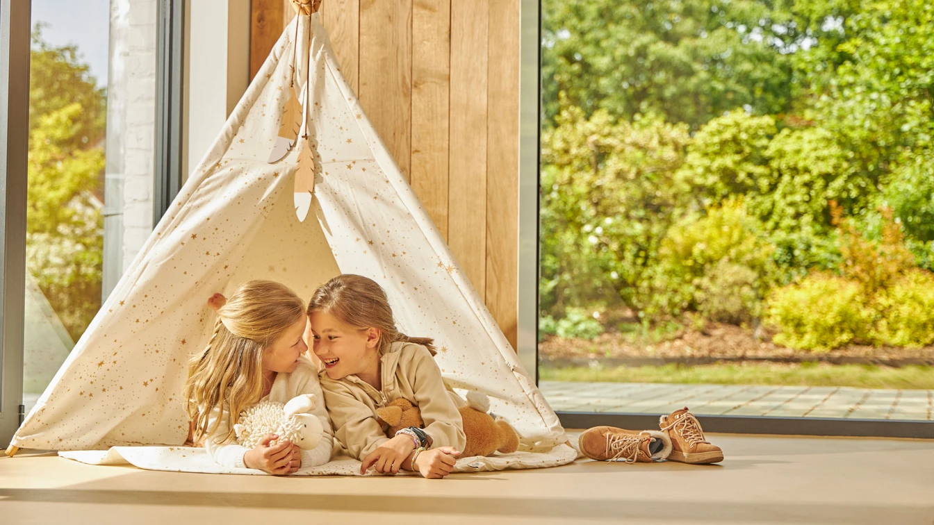 Two young girls smiling and lying inside a cream star-patterned play tent near a large window with green garden outside.