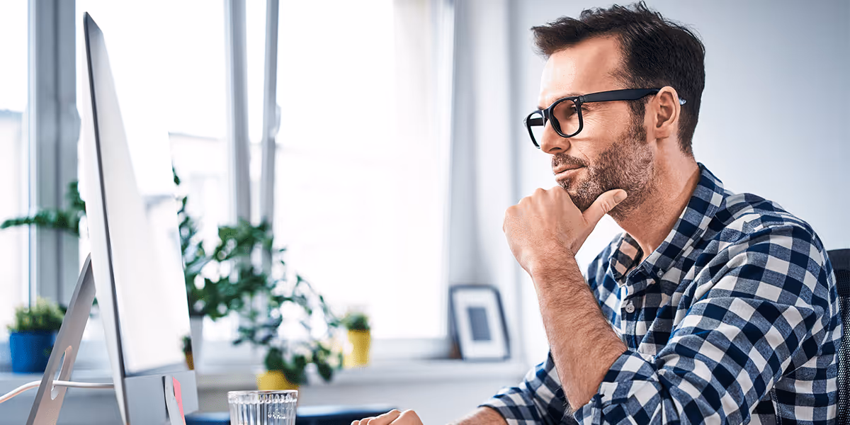 Man wearing glasses and a checkered shirt works thoughtfully at a desktop computer in a bright office.