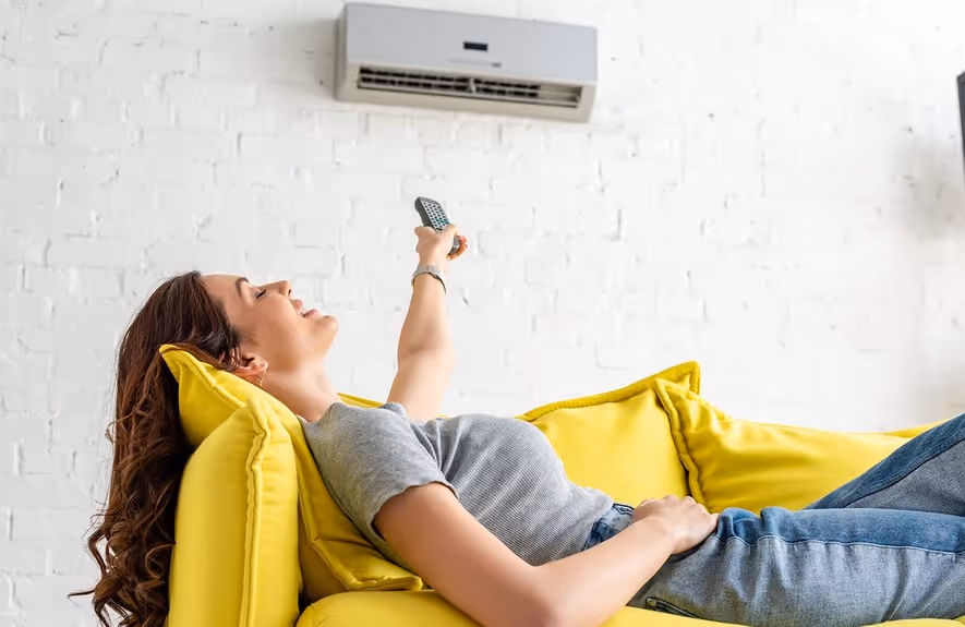 Woman reclining on a yellow couch pointing a remote control at a wall-mounted air conditioner.