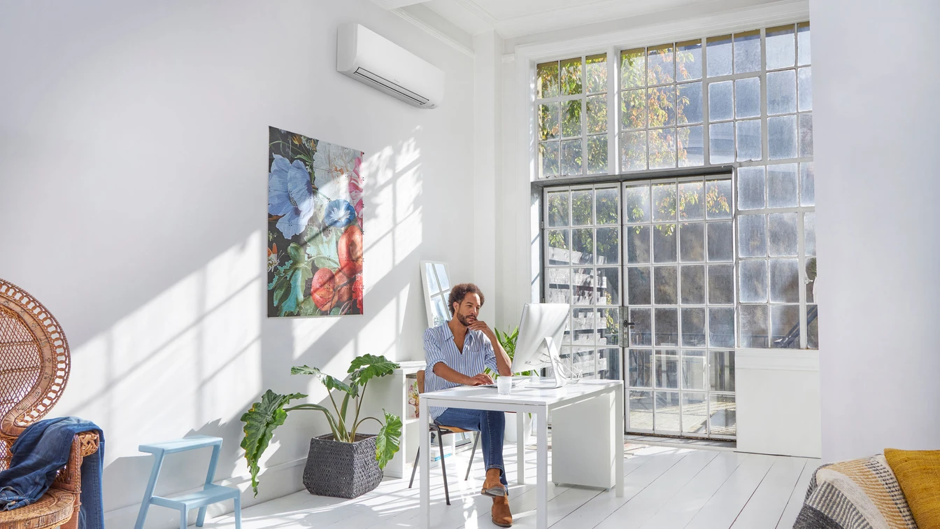 Man sitting at a white desk in a bright, airy room with large grid windows, working on a desktop computer.