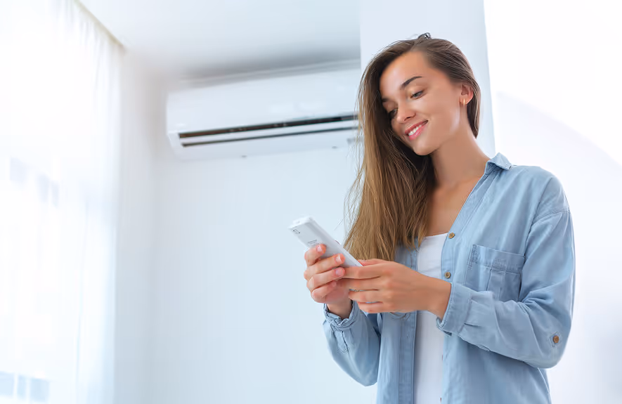 Woman holding remote control, smiling, with wall-mounted air conditioner in the background.