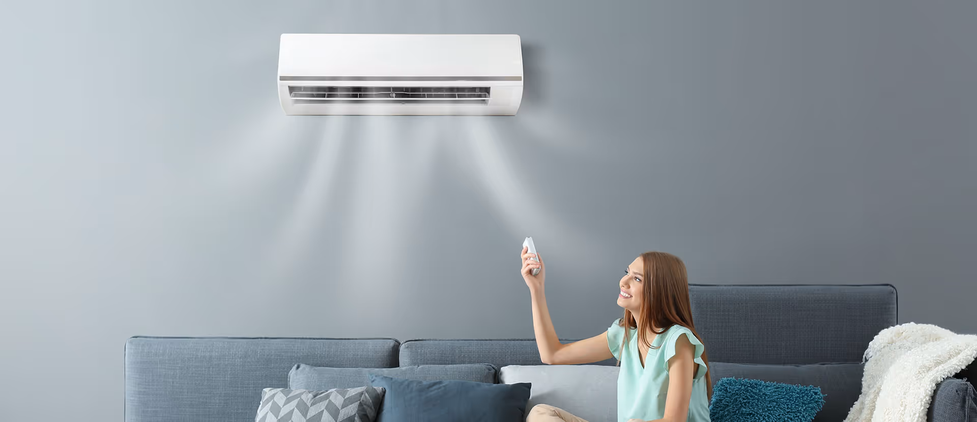 Smiling woman sitting on a sofa using remote to control wall-mounted air conditioner blowing cool air.
