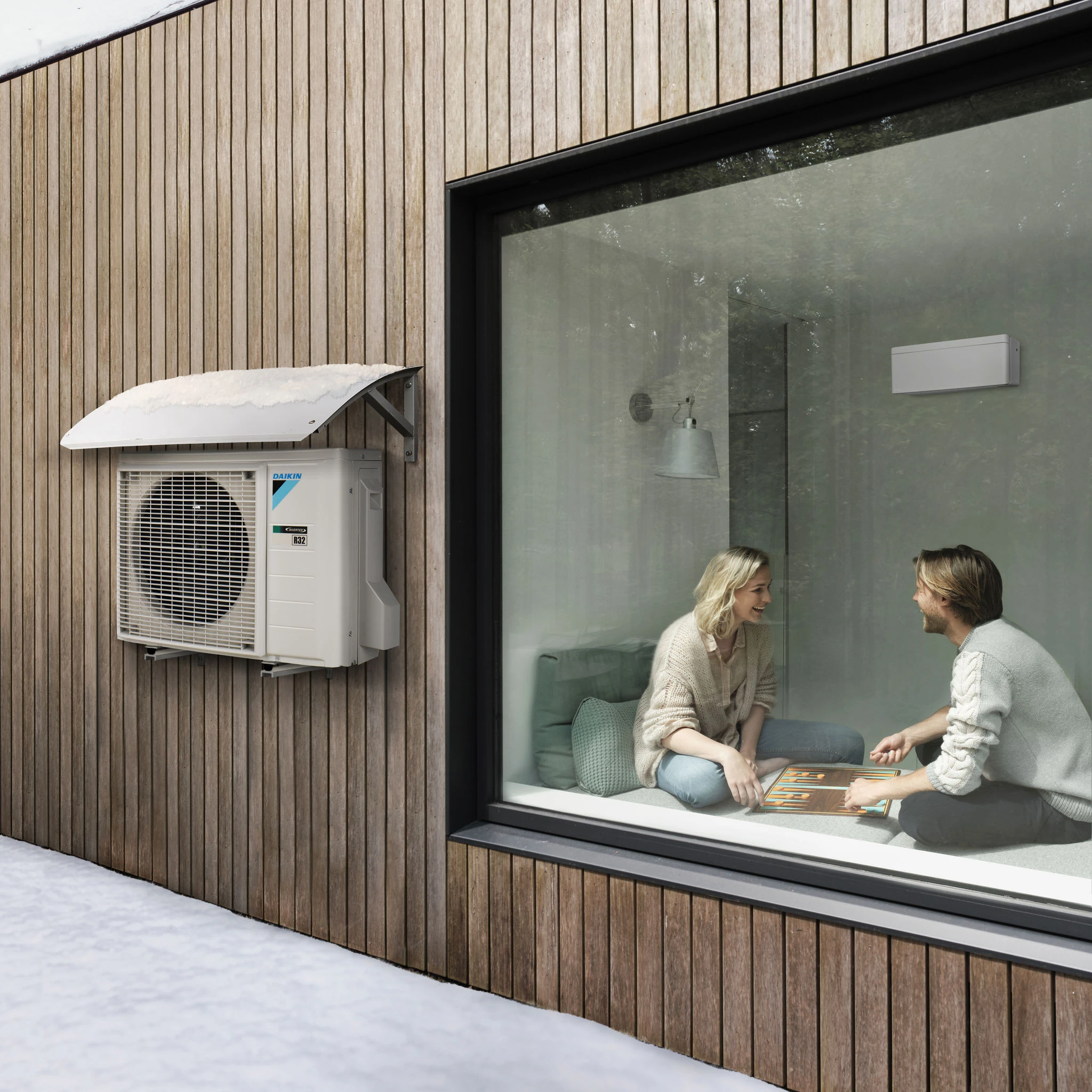 Exterior wooden wall with a snow-covered Daikin air conditioning unit next to a large window showing a man and woman playing backgammon inside a cozy room.