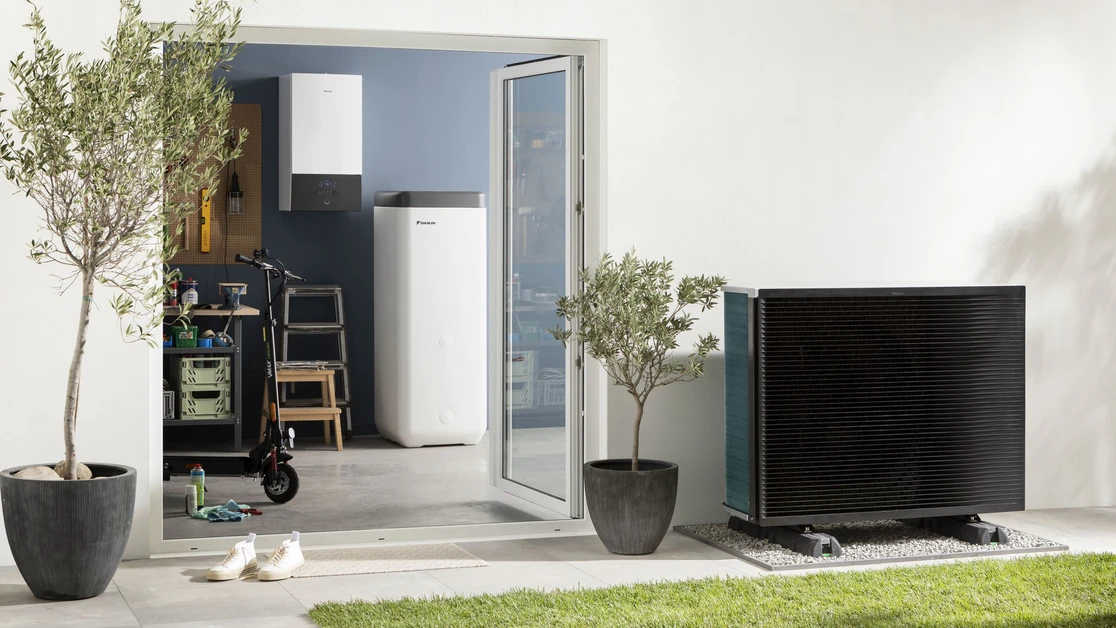 Outdoor view of a modern heat pump unit on a gravel bed next to two potted plants, with a partially open glass door leading to a garage containing a scooter, ladder, and white storage units.