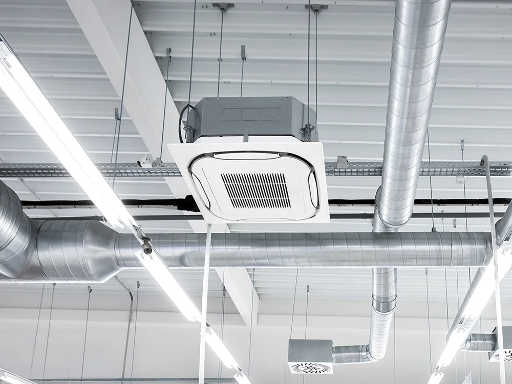 Plafond industriel avec conduits d'aération en métal, éclairage fluorescent suspendu et un système de climatisation encastré.