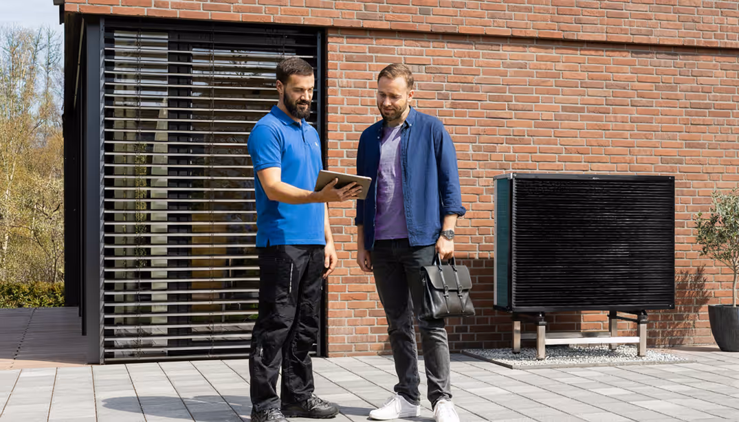 Deux hommes debout devant une maison en briques, l'un montre quelque chose sur une tablette à l'autre.