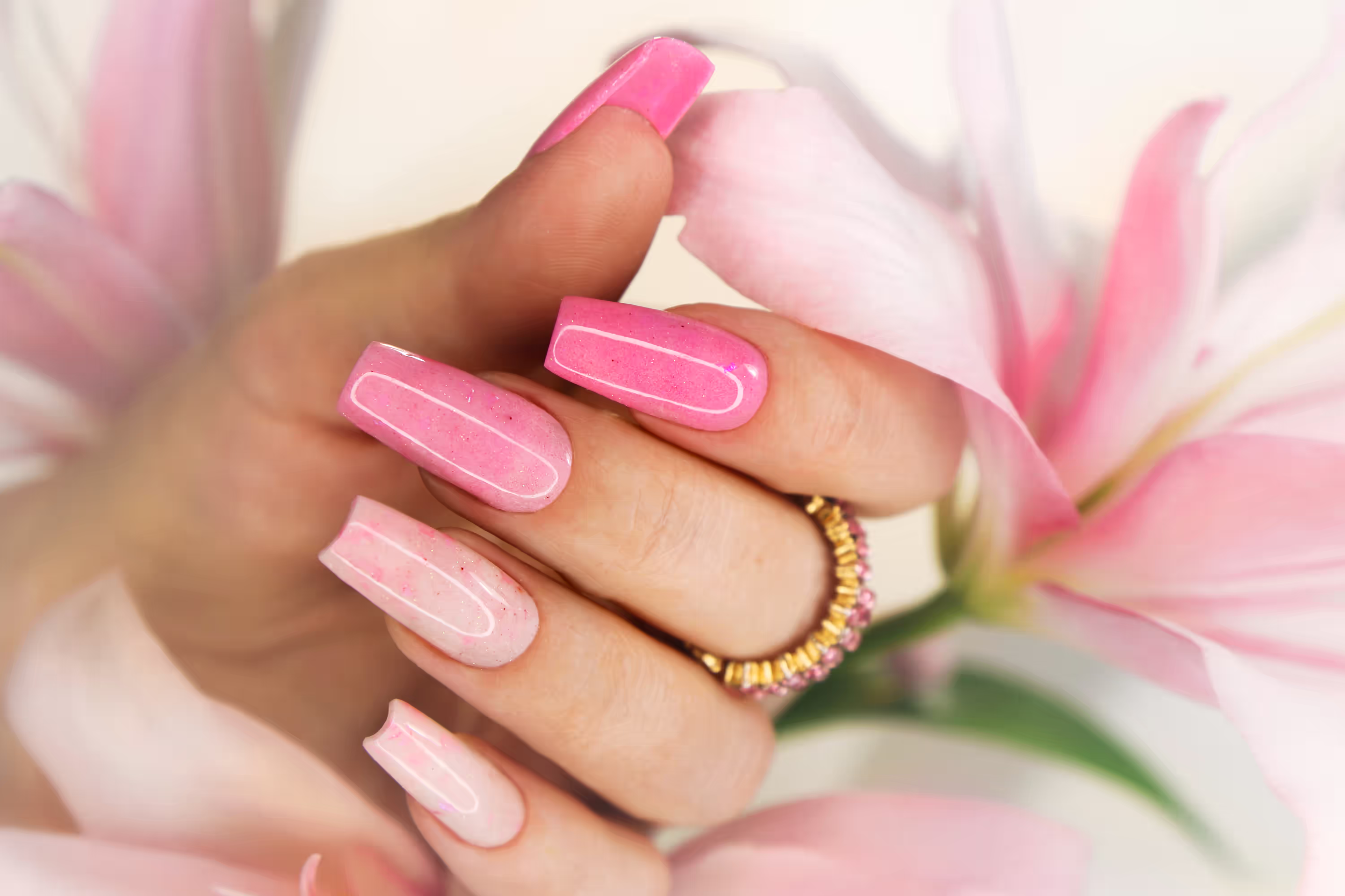 Close-up of a hand with glossy pink bio gel nails in an ombre design, accessorized with a gold ring and surrounded by soft pink flower petals.
