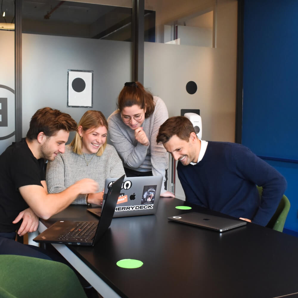 Four young adults smiling and looking at laptops around a black table in a modern office.