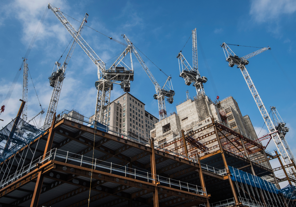 Construction site with steel framework and multiple tower cranes against a blue sky.