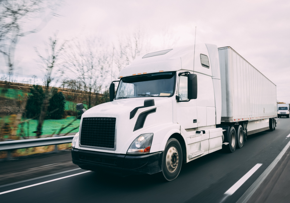 White semi-truck with trailer driving on highway with blurred trees and greenery in the background.