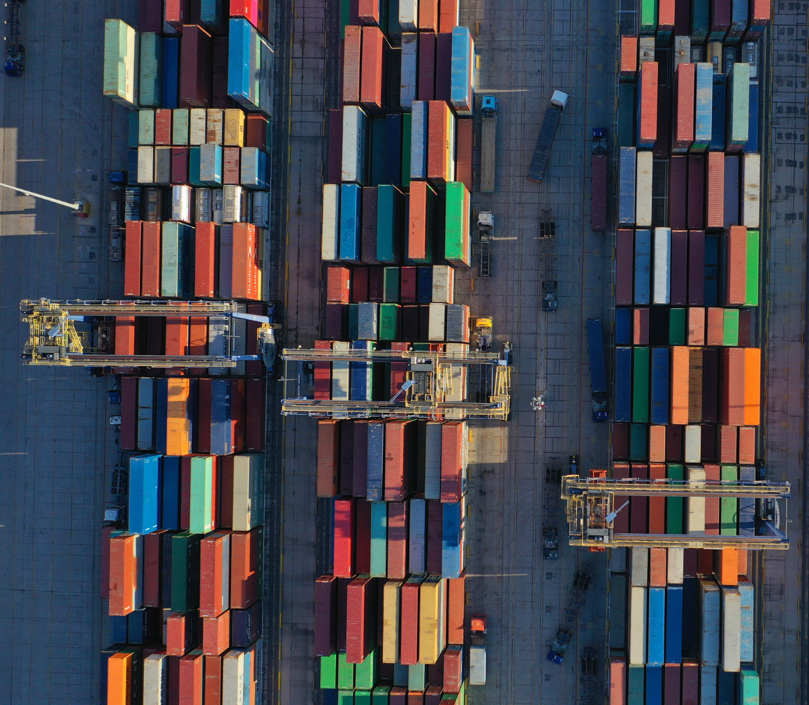 Aerial view of colorful shipping containers neatly arranged in rows at a busy port with container cranes.