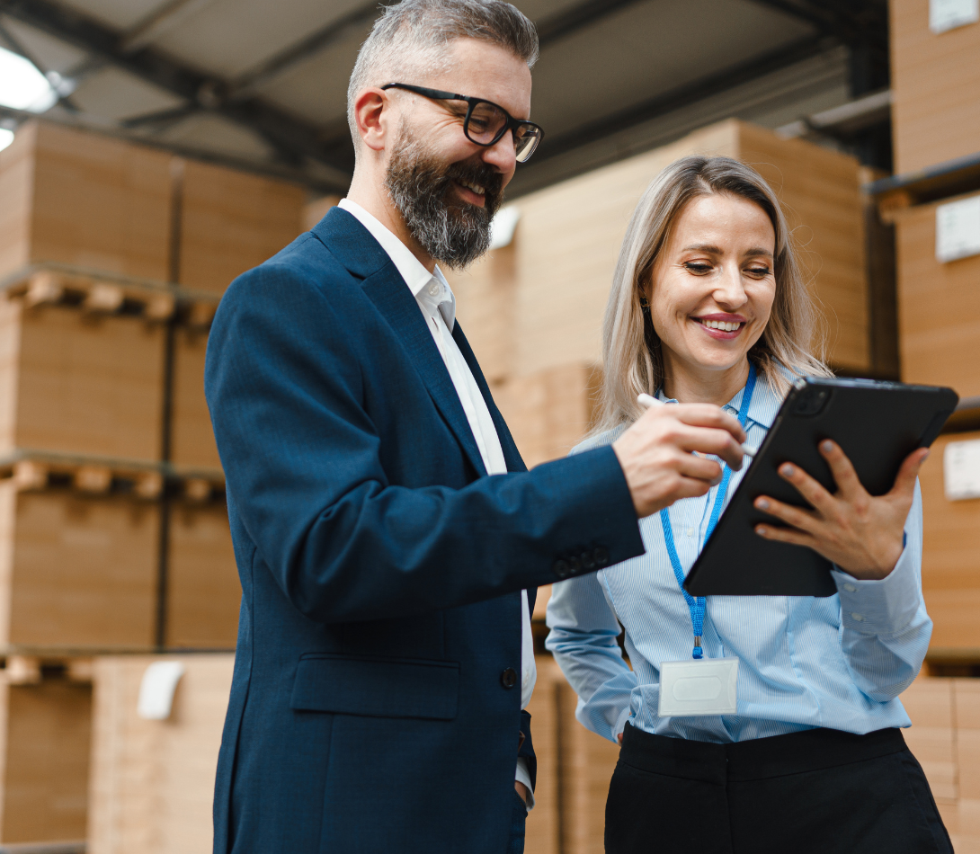 Two warehouse employees smiling while reviewing information on a digital tablet in a storage area with stacked cardboard boxes.